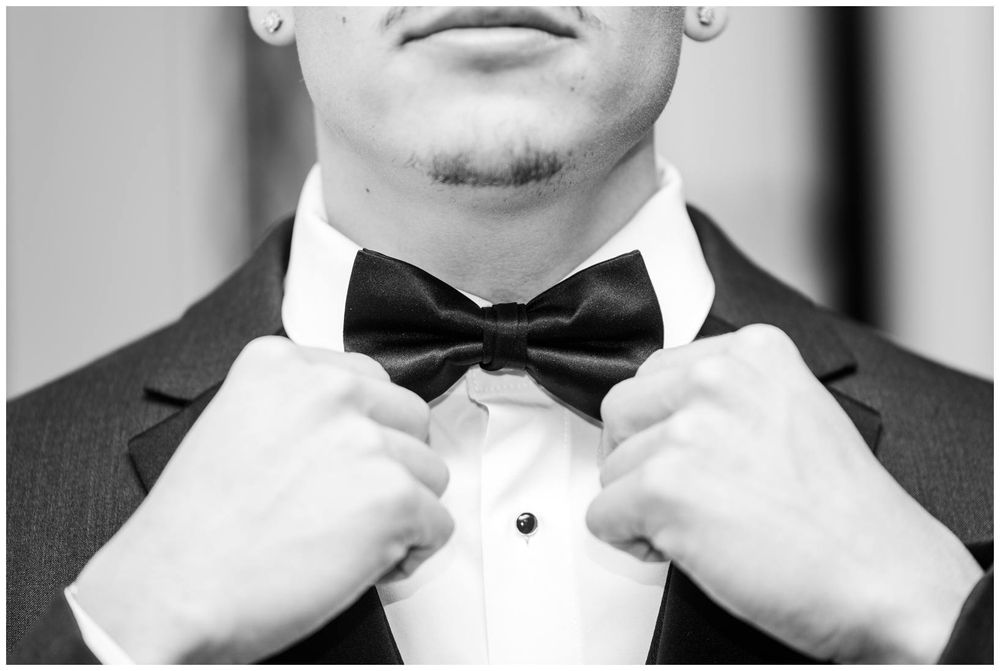 Groom in tuxedo adjusting black bow tie, close-up black and white wedding photo.