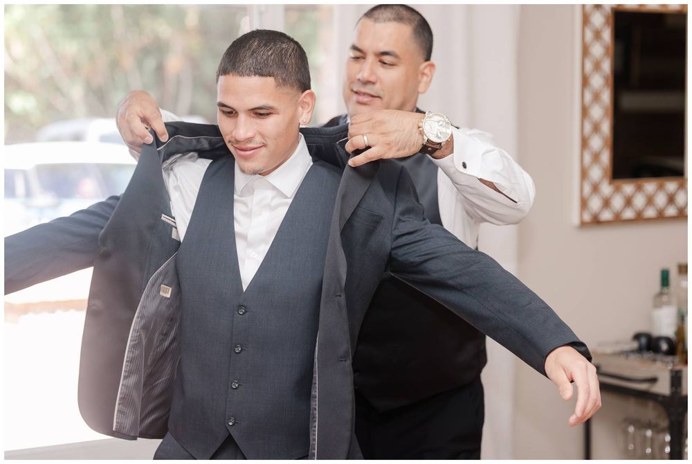 Groom being helped into his grey suit jacket by a groomsman before the wedding ceremony.