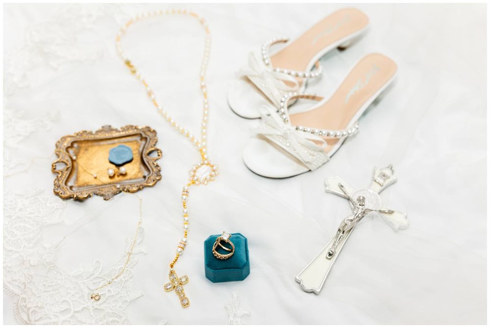 Bridal flat sandals with pearl details beside a rosary, crucifix, ring box, and ornate picture frame on white lace.
