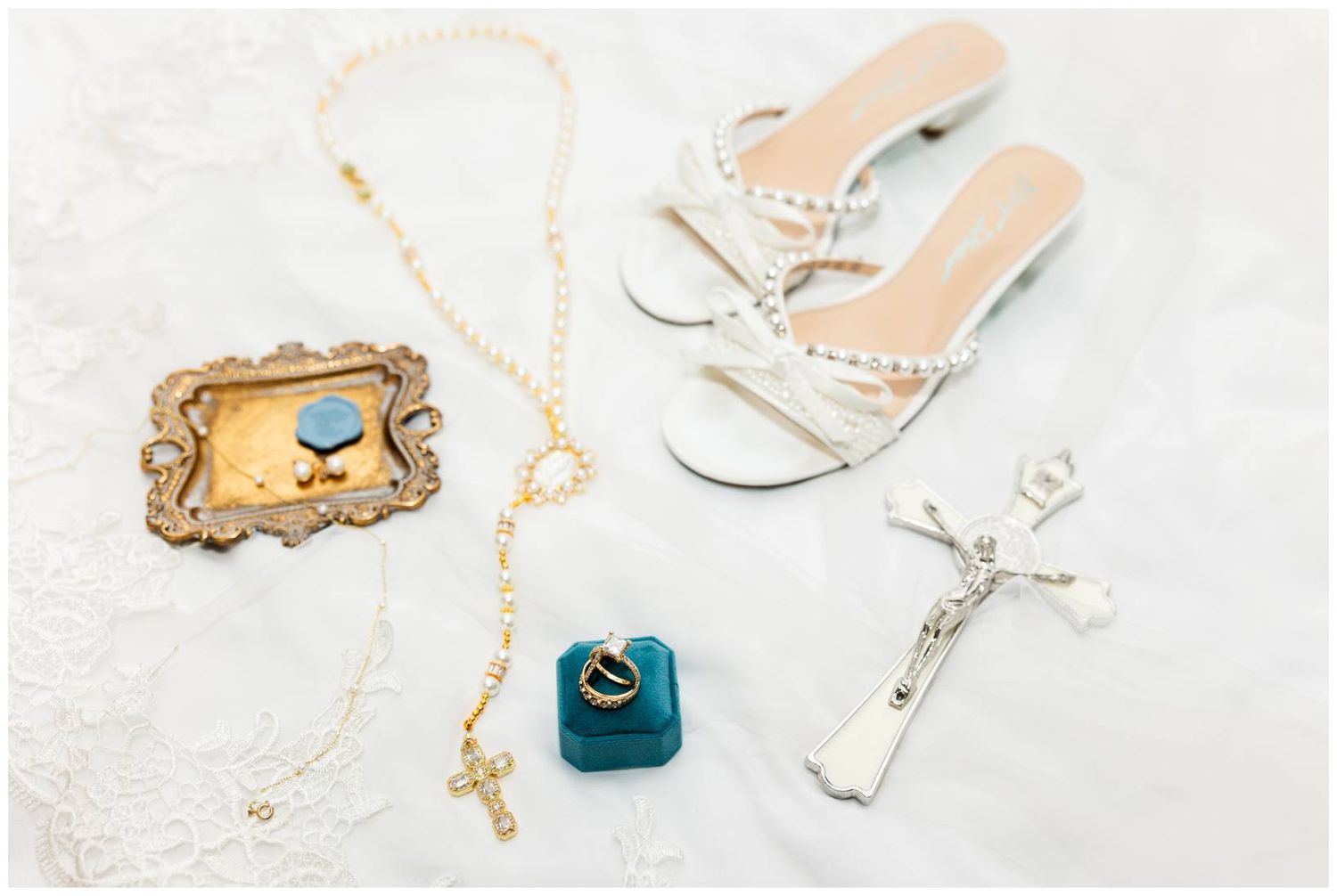 Bridal flat sandals with pearl details beside a rosary, crucifix, ring box, and ornate picture frame on white lace.