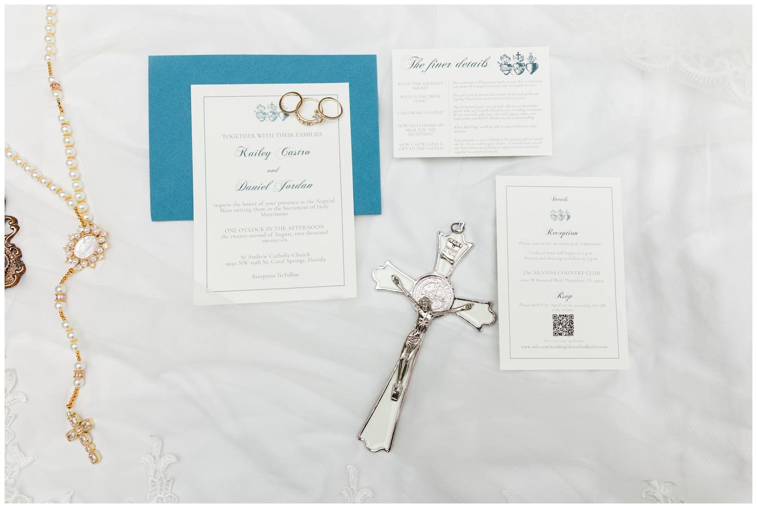 Wedding invitation suite with teal accents, cross, rosary beads, and stationery cards displayed on white lace fabric.