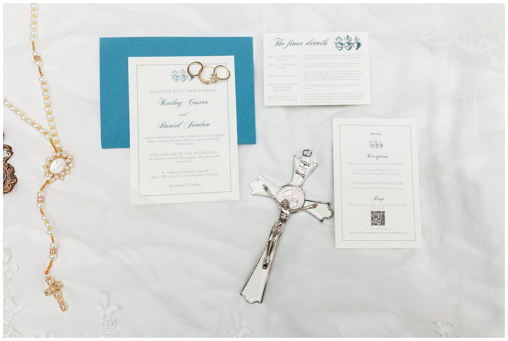 Wedding invitation suite with teal accents, cross, rosary beads, and stationery cards displayed on white lace fabric.