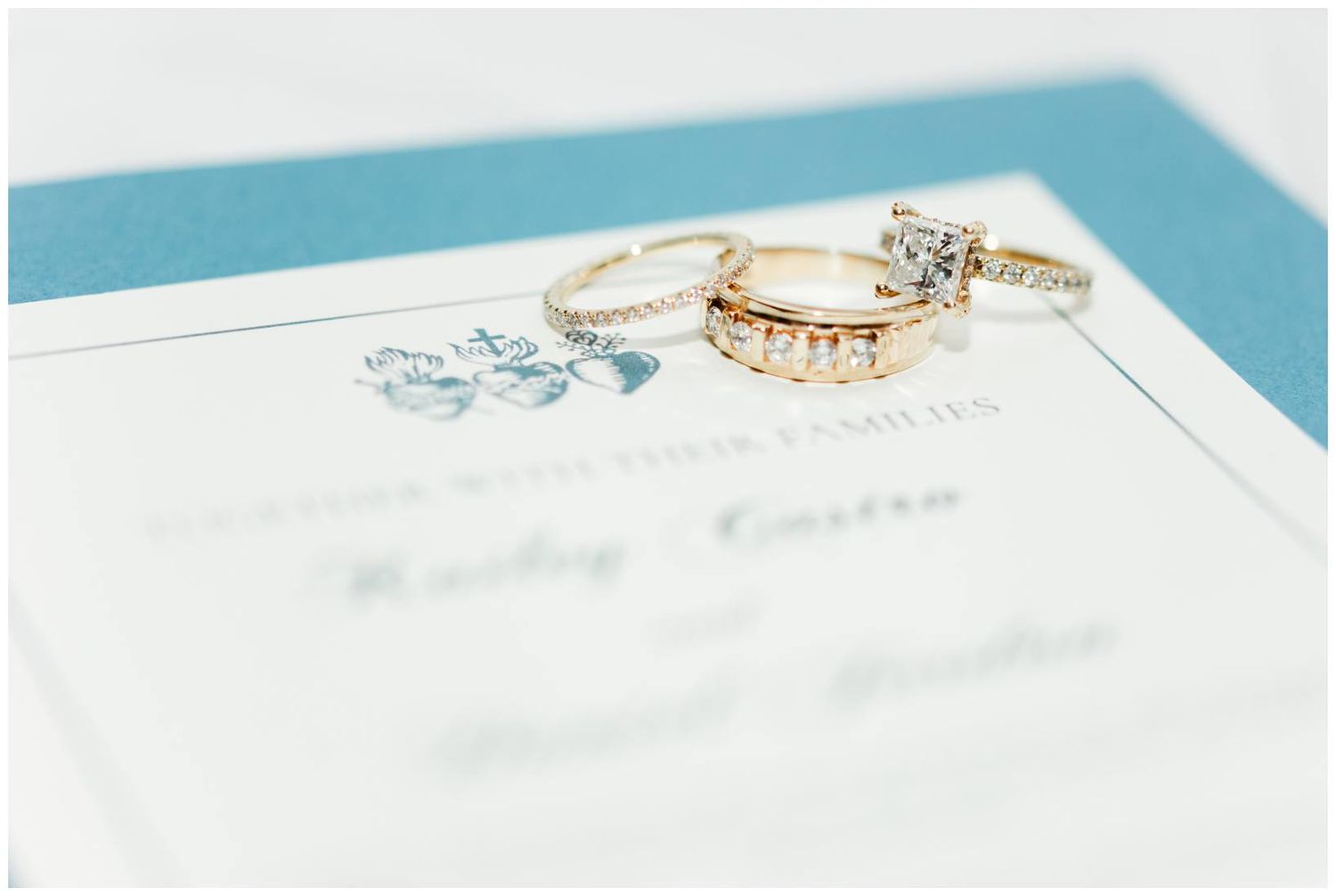 Gold wedding rings and diamond engagement ring displayed on a teal and white wedding invitation.