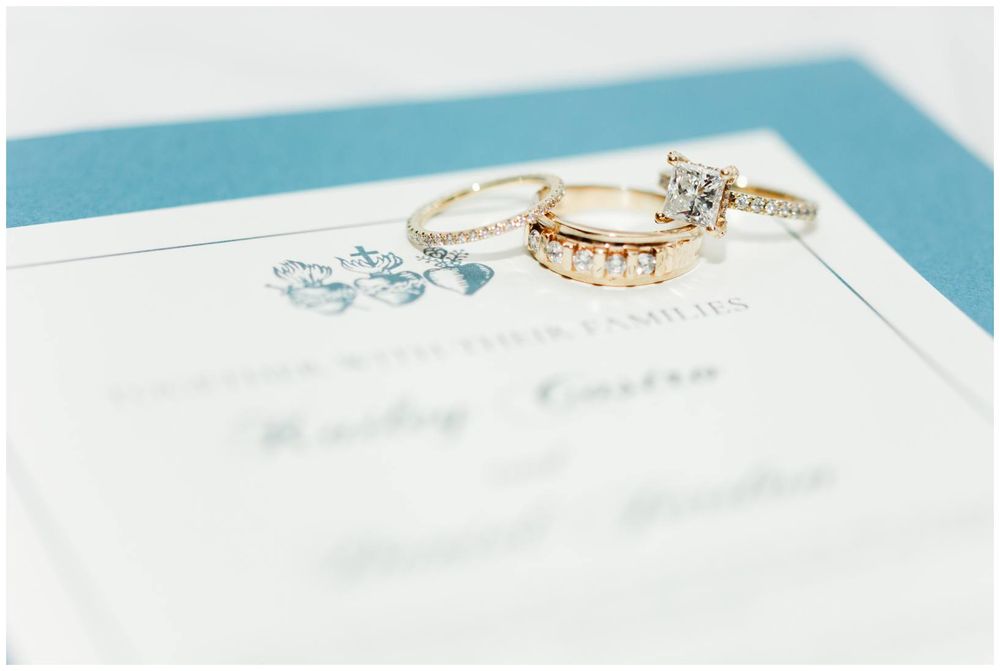 Gold wedding rings and diamond engagement ring displayed on a teal and white wedding invitation.