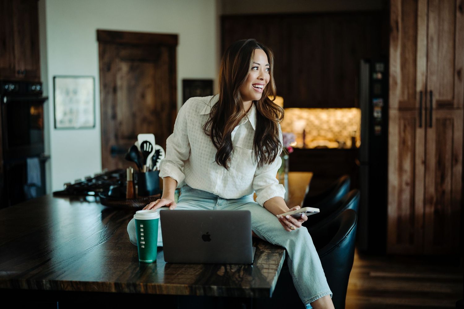 Woman smiling while sitting on desk with MacBook laptop and phone in modern home office setting.