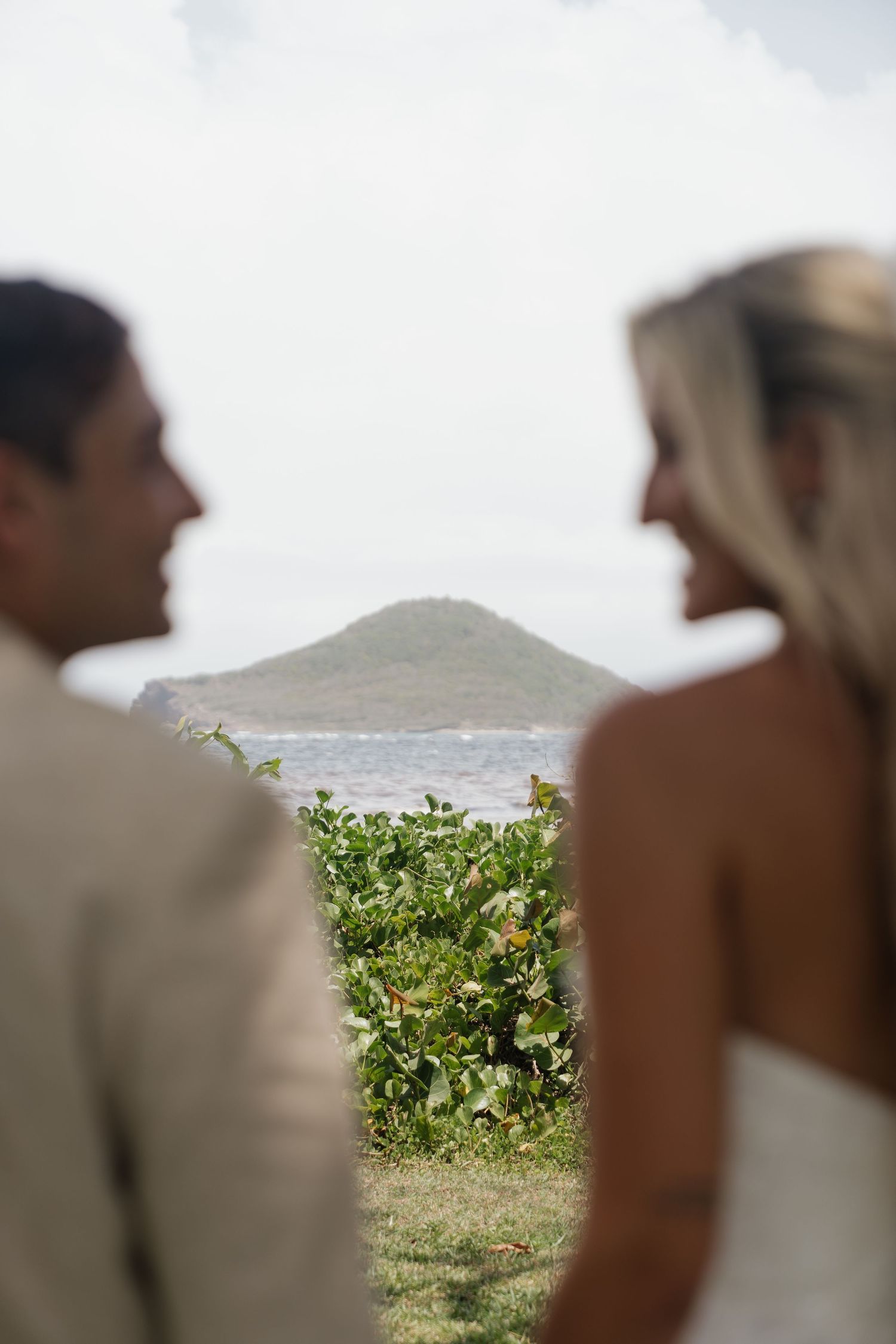 Couple in wedding attire facing each other with tropical island and ocean view in background.