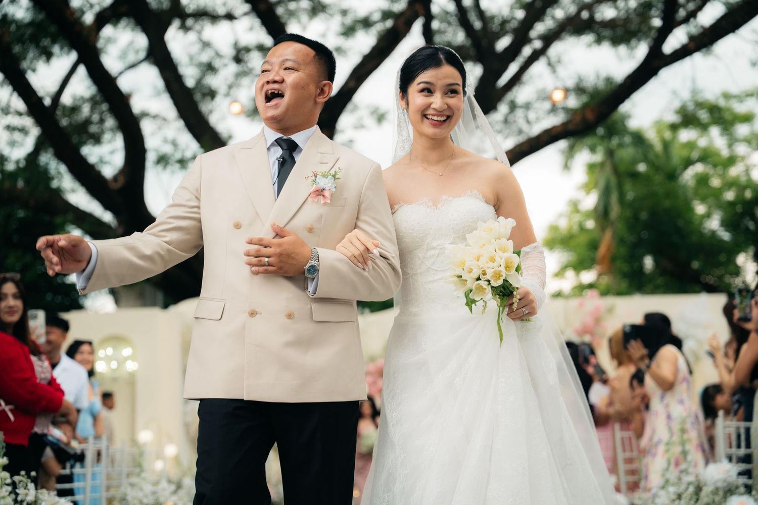 Joyful newlywed couple walking down outdoor wedding aisle, groom in beige suit, bride in white strapless gown holding bouquet.