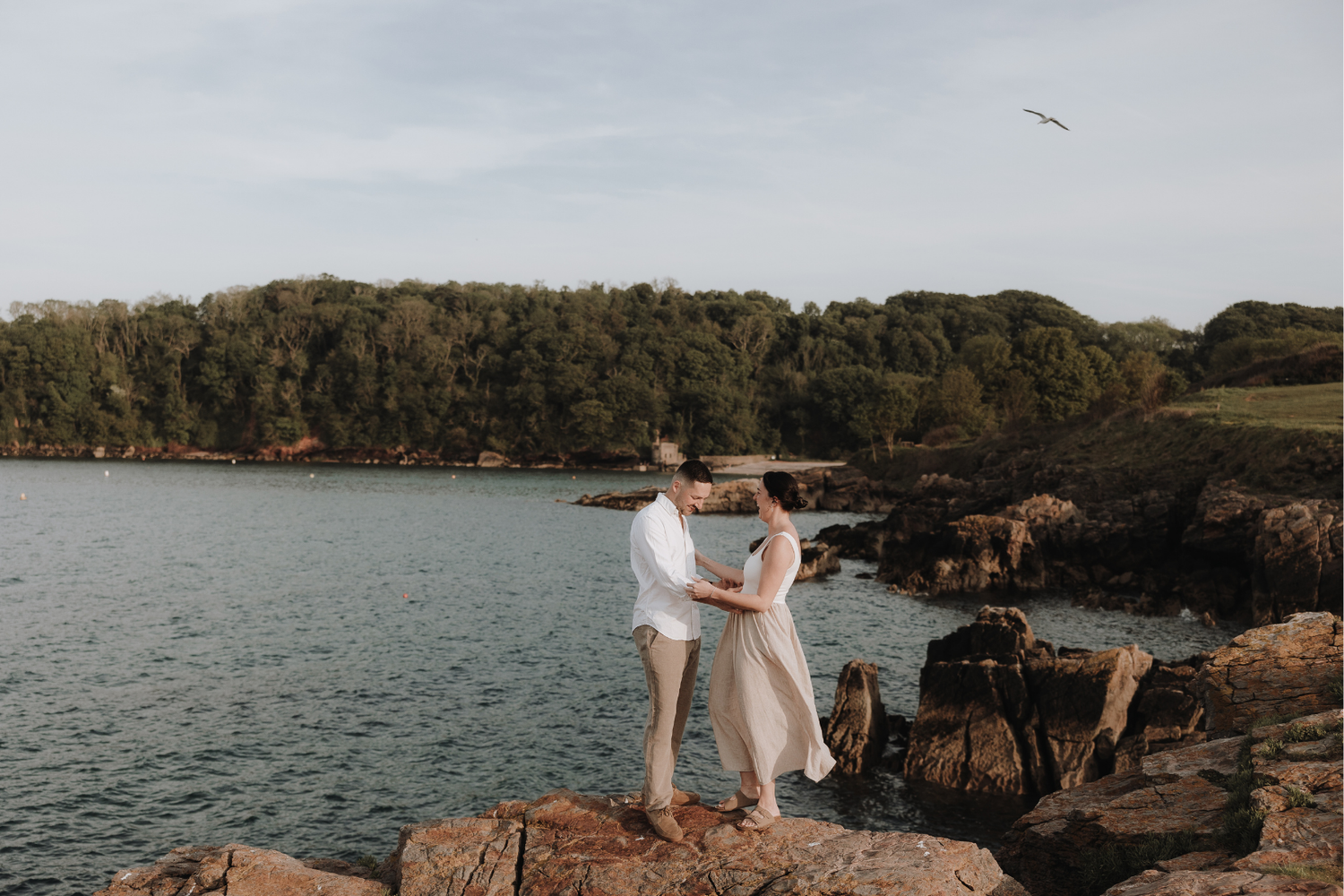 Couple holding hands on rocky coastal shore with lush green trees and calm blue water in background.