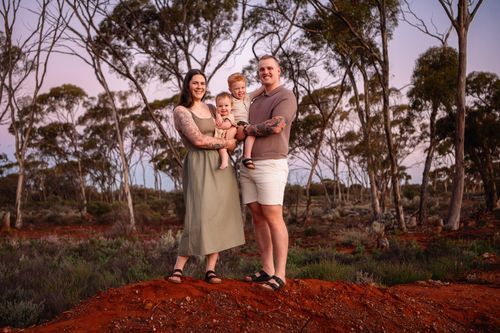Family of four poses on red dirt path surrounded by Australian bush trees at golden hour sunset.