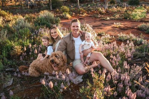 Family with two kids and a golden doodle sitting among pink wildflowers on red dirt at golden hour.