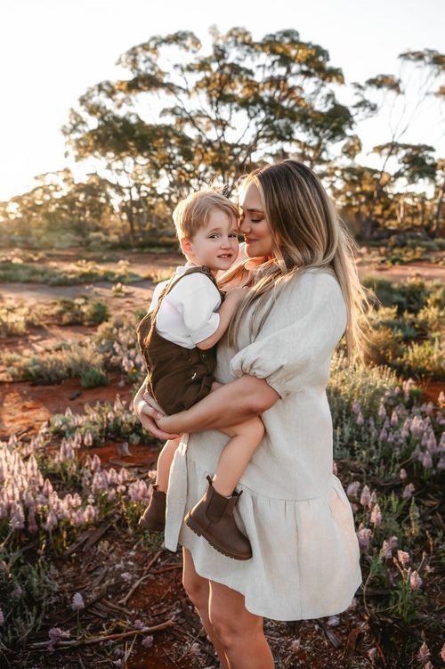 A mother tenderly kisses her toddler son while holding him in a sunlit wildflower field.