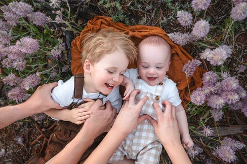 Two laughing babies lying on an orange blanket surrounded by purple wildflowers, held by adult hands outdoors.