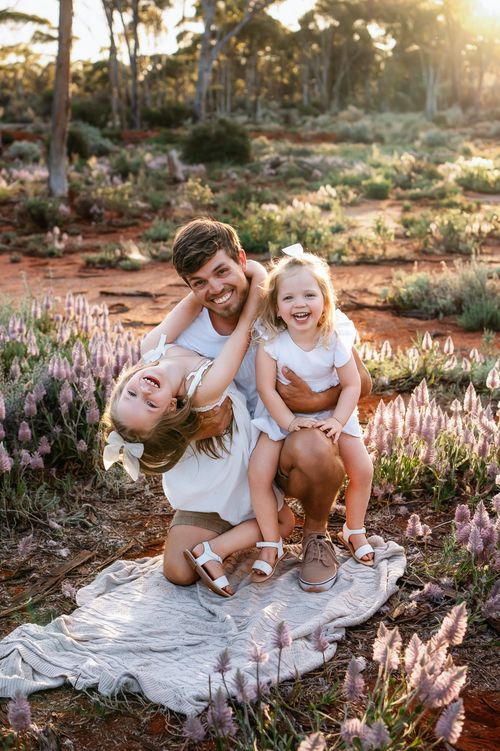 A smiling father plays with his two laughing daughters in a blooming wildflower meadow at sunset.