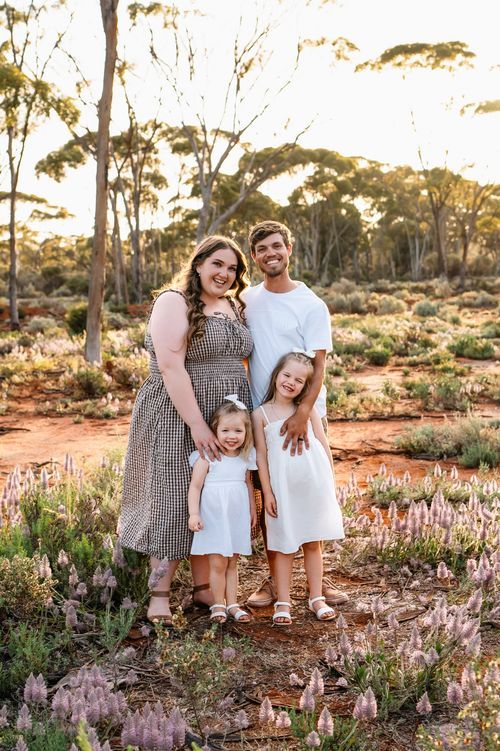 Family of four posing outdoors in Australian bushland with red dirt and wildflowers, wearing white and grey outfits.