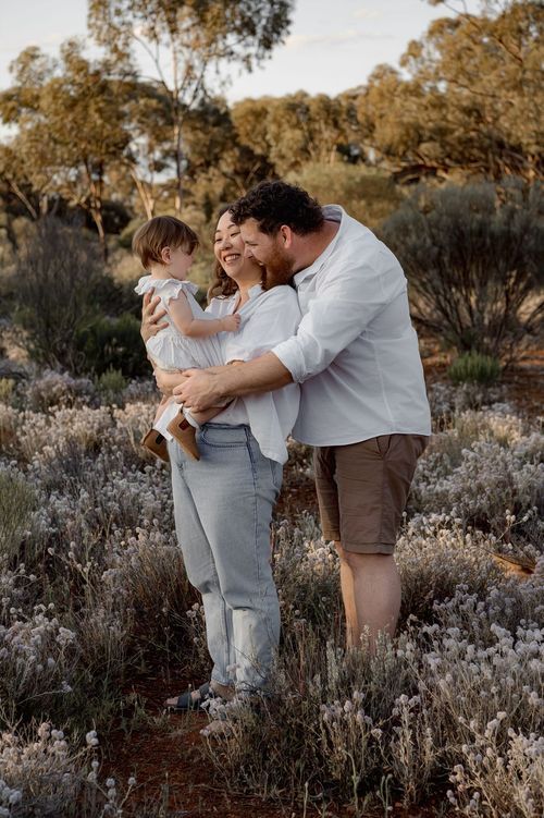 Happy family with baby enjoying golden hour in Australian bush landscape, parents smiling and embracing child.