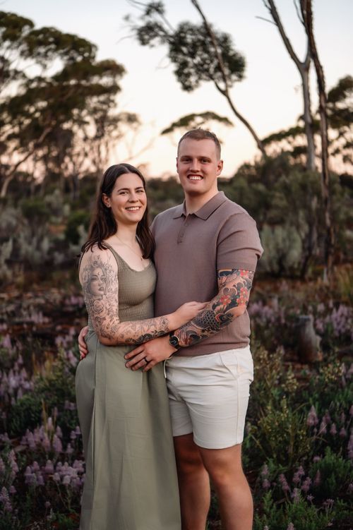 Couple with tattoos smiling outdoors at sunset, surrounded by wildflowers and eucalyptus trees.