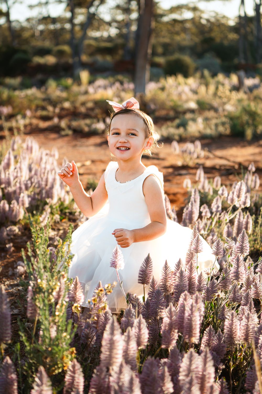 Toddler girl in white tutu dress and bow headband smiling among purple wildflowers in a sunlit field.