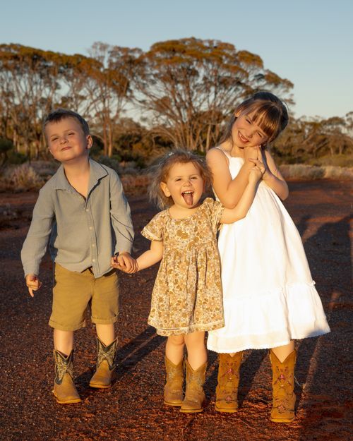 Three young children in cowboy boots hold hands on a red dirt road at golden hour in the Australian outback.