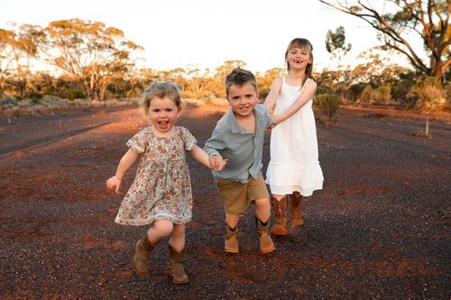 Three happy children playing on a red dirt path surrounded by Australian outback trees at golden hour.