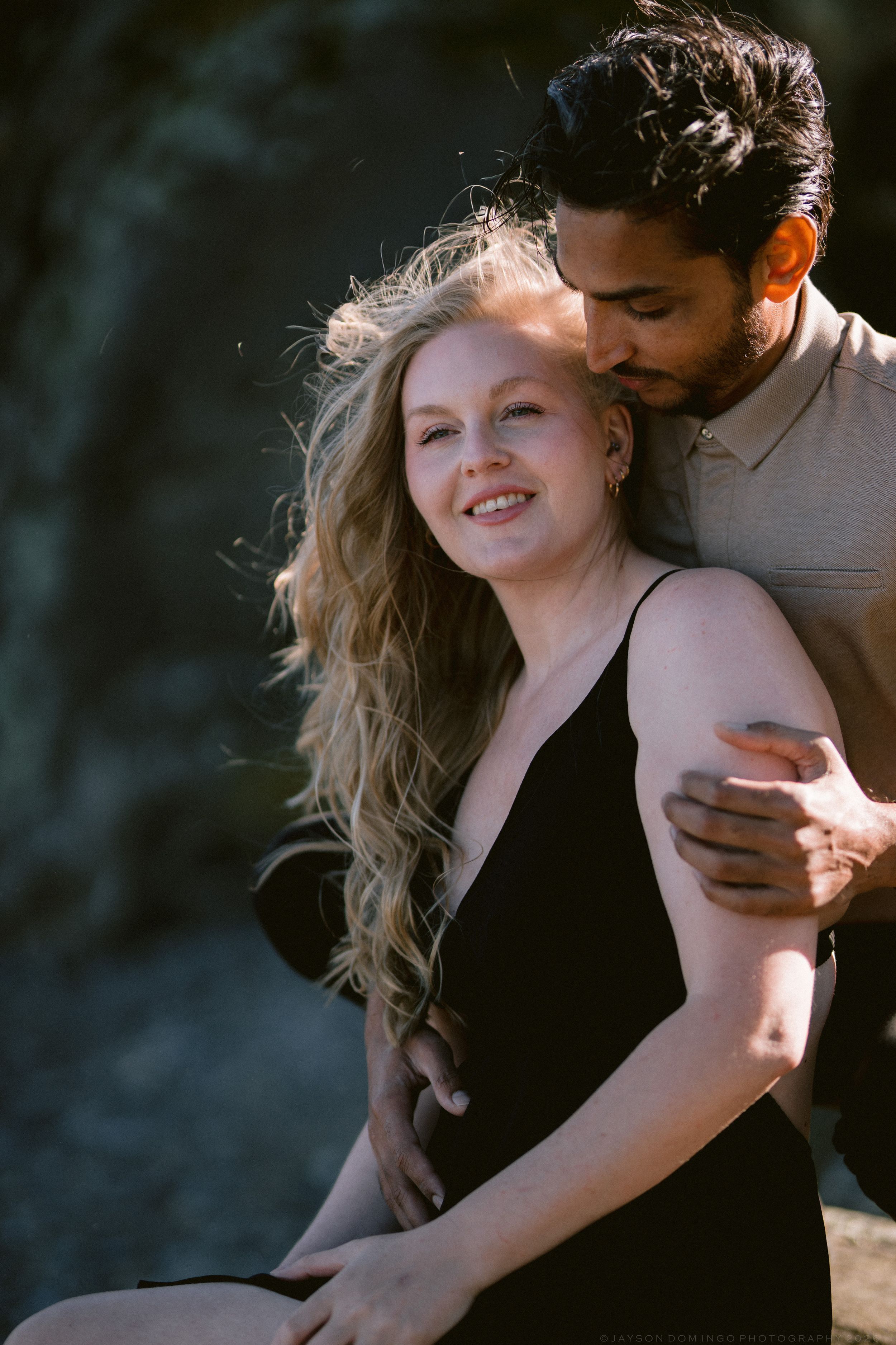 Man tenderly kissing woman's head while embracing her outdoors in warm golden light.