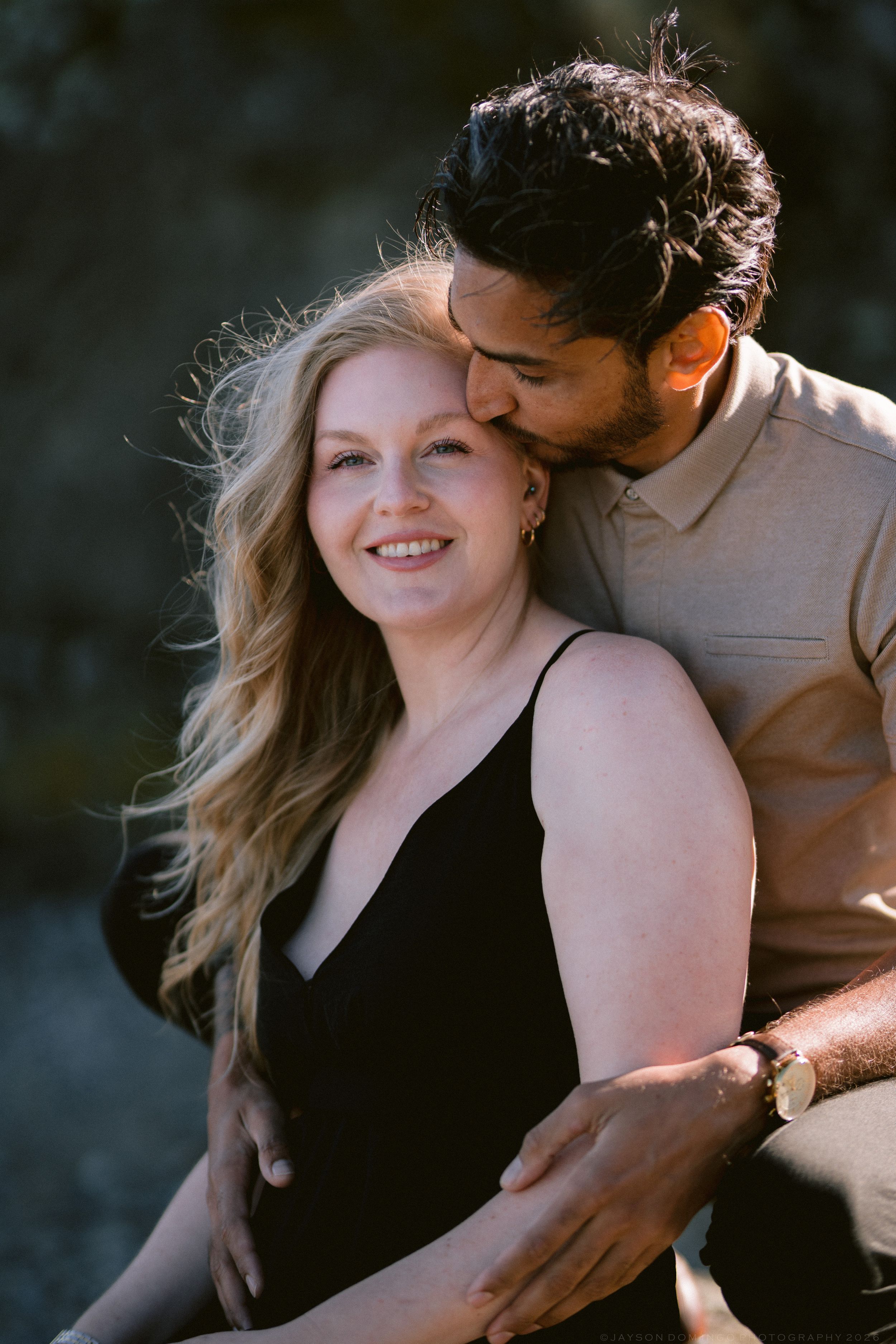 A couple shares a tender moment outdoors, man kissing womans temple while she smiles warmly in a black dress.