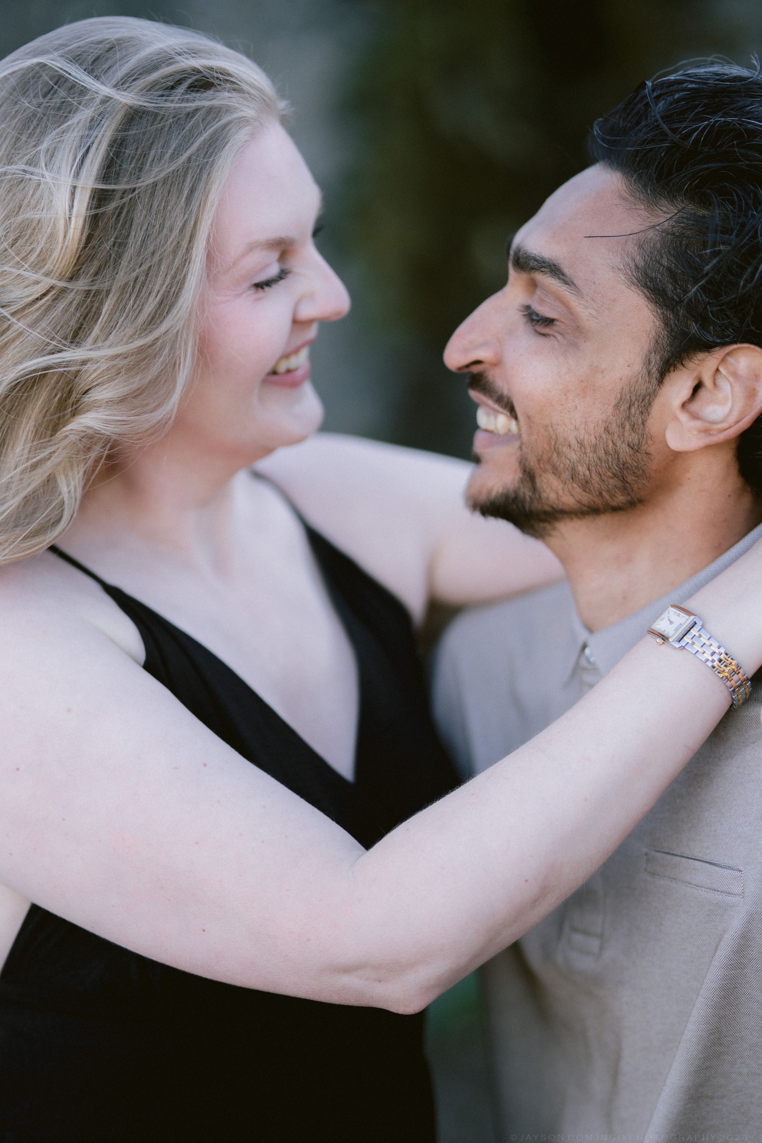 A smiling couple gazes at each other, woman in black dress with hand on man's shoulder in beige suit.