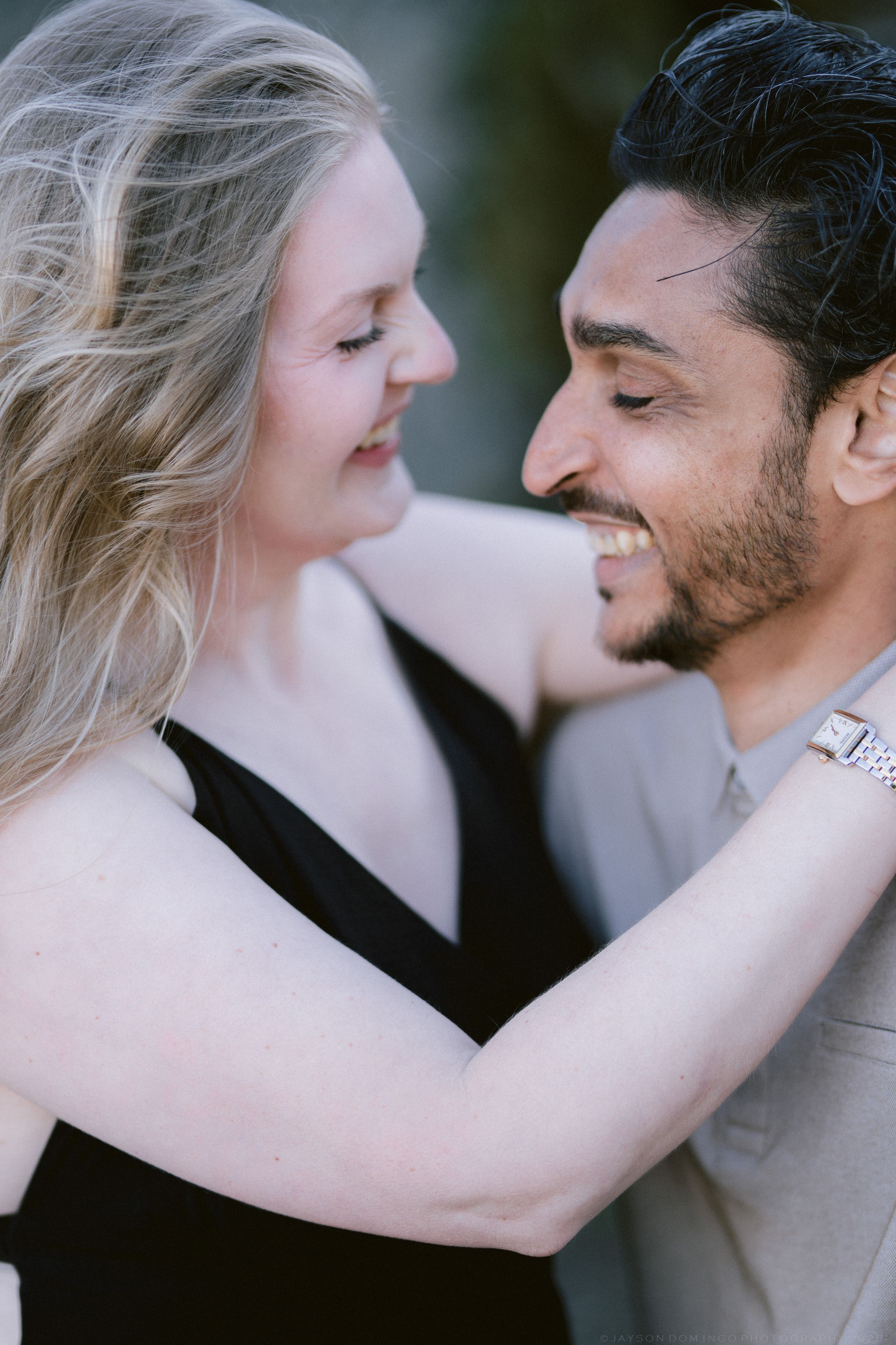 A couple sharing a romantic moment, woman in black dress with arms around a smiling bearded man in a gray suit.