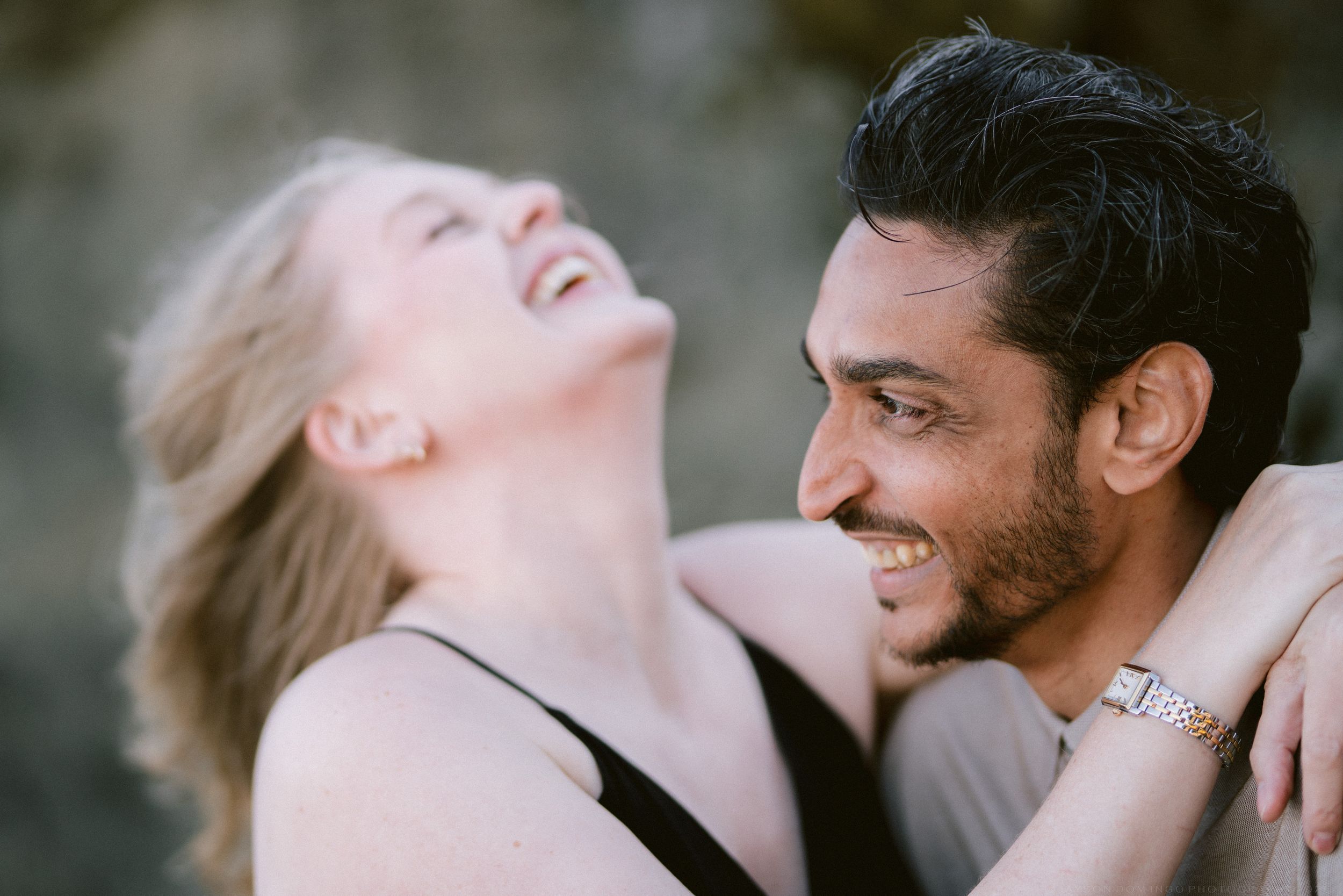 A laughing blonde woman and smiling dark-haired man share a joyful moment together outdoors.