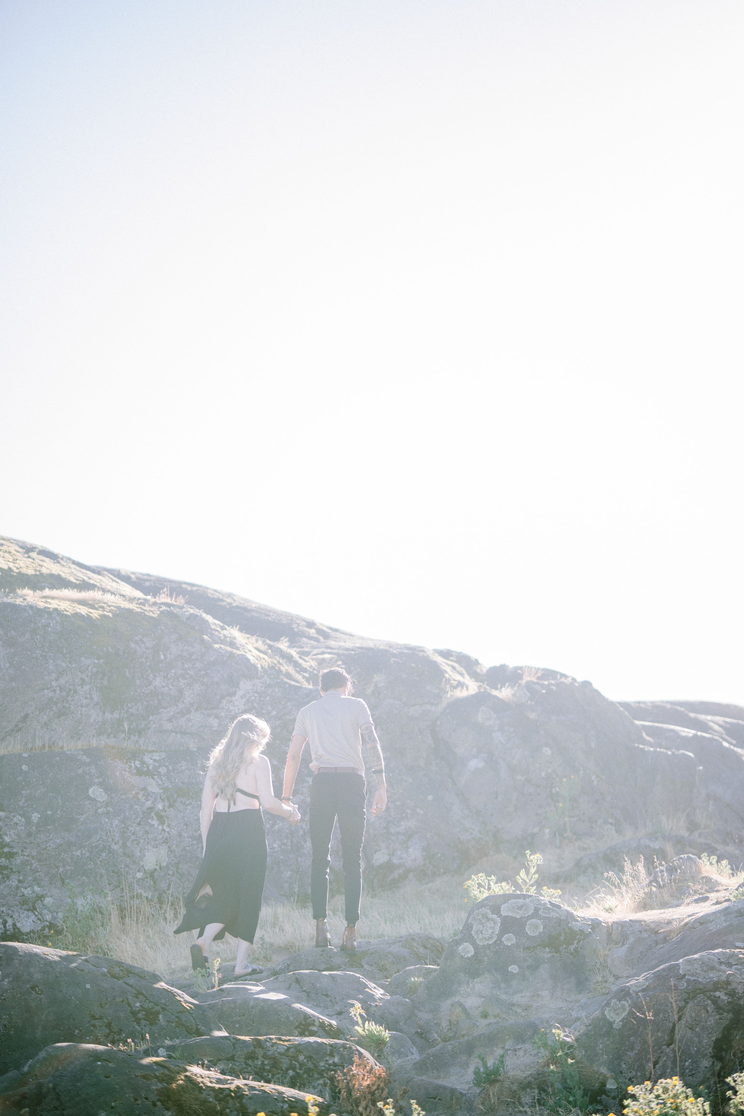 A couple holds hands walking toward sunlit rocky hills, photographed from behind in bright glowing backlight.