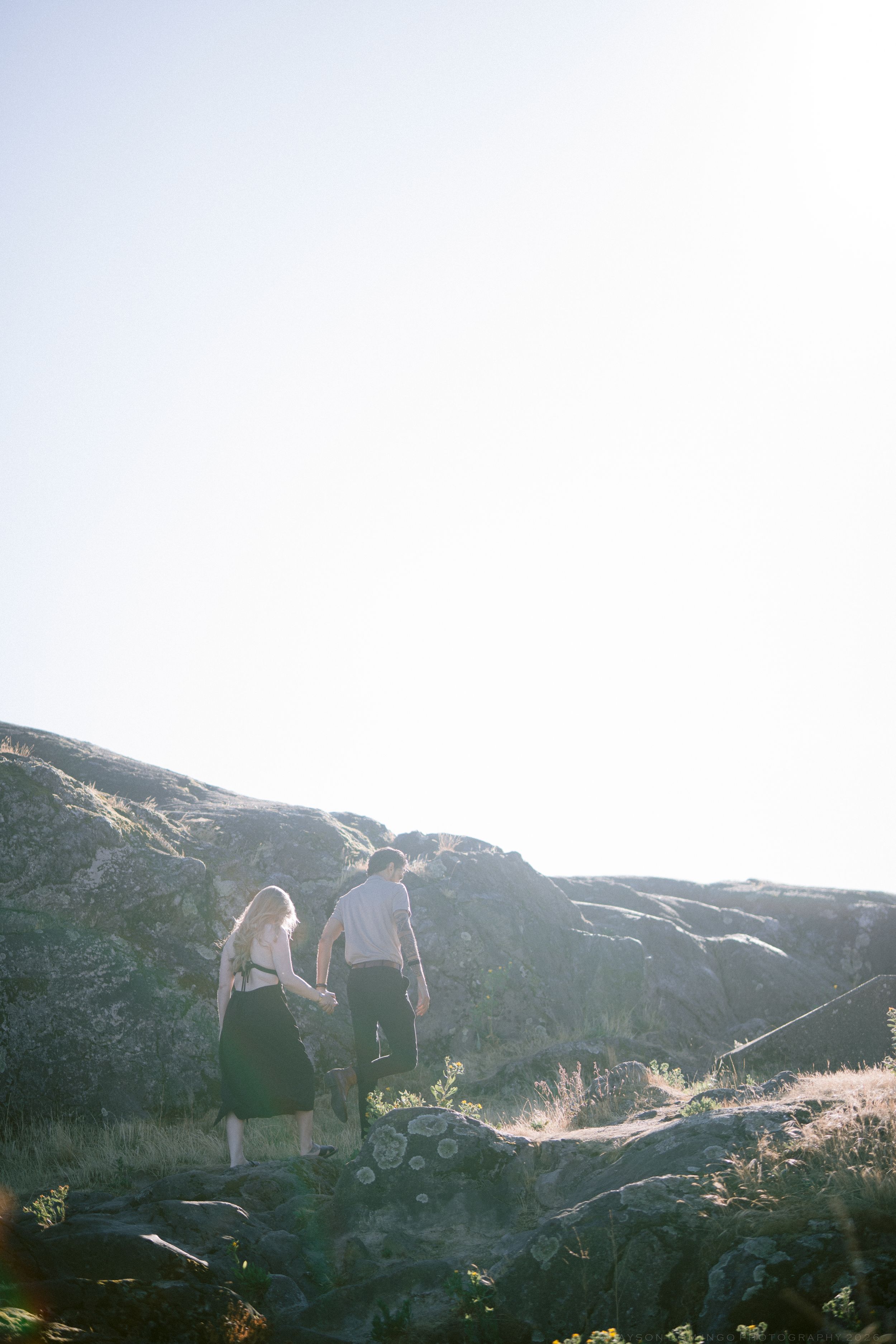 Couple holding hands walking on rocky landscape silhouetted against bright clear sky.