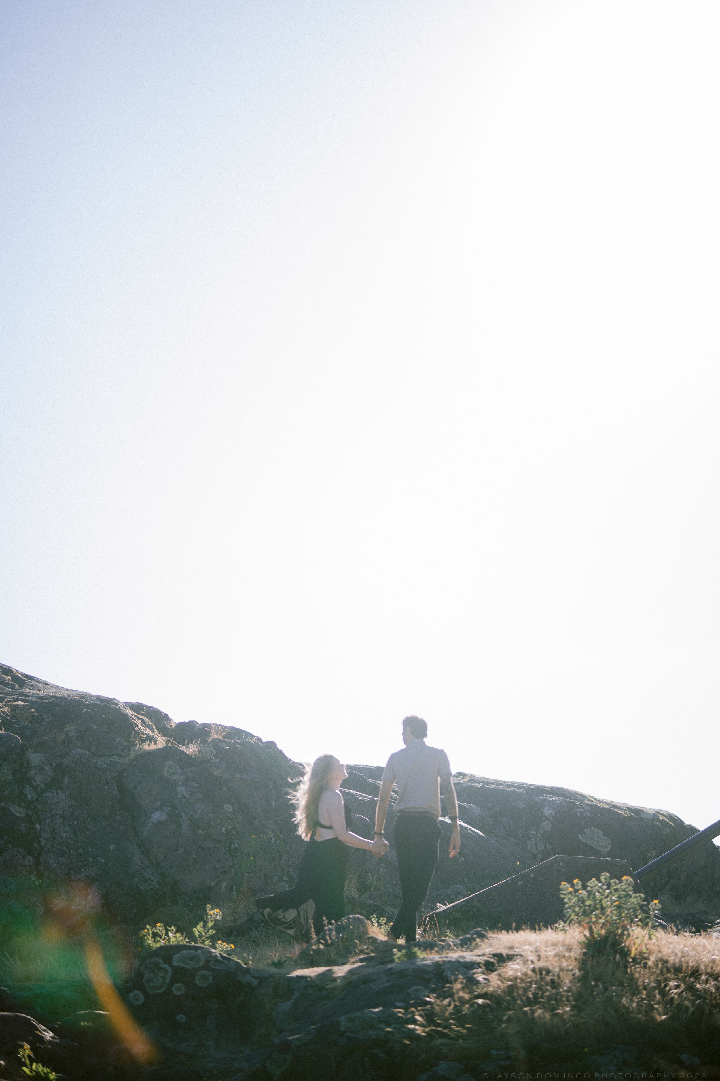 Couple hiking on rocky terrain with bright sunlight and clear sky in the background.