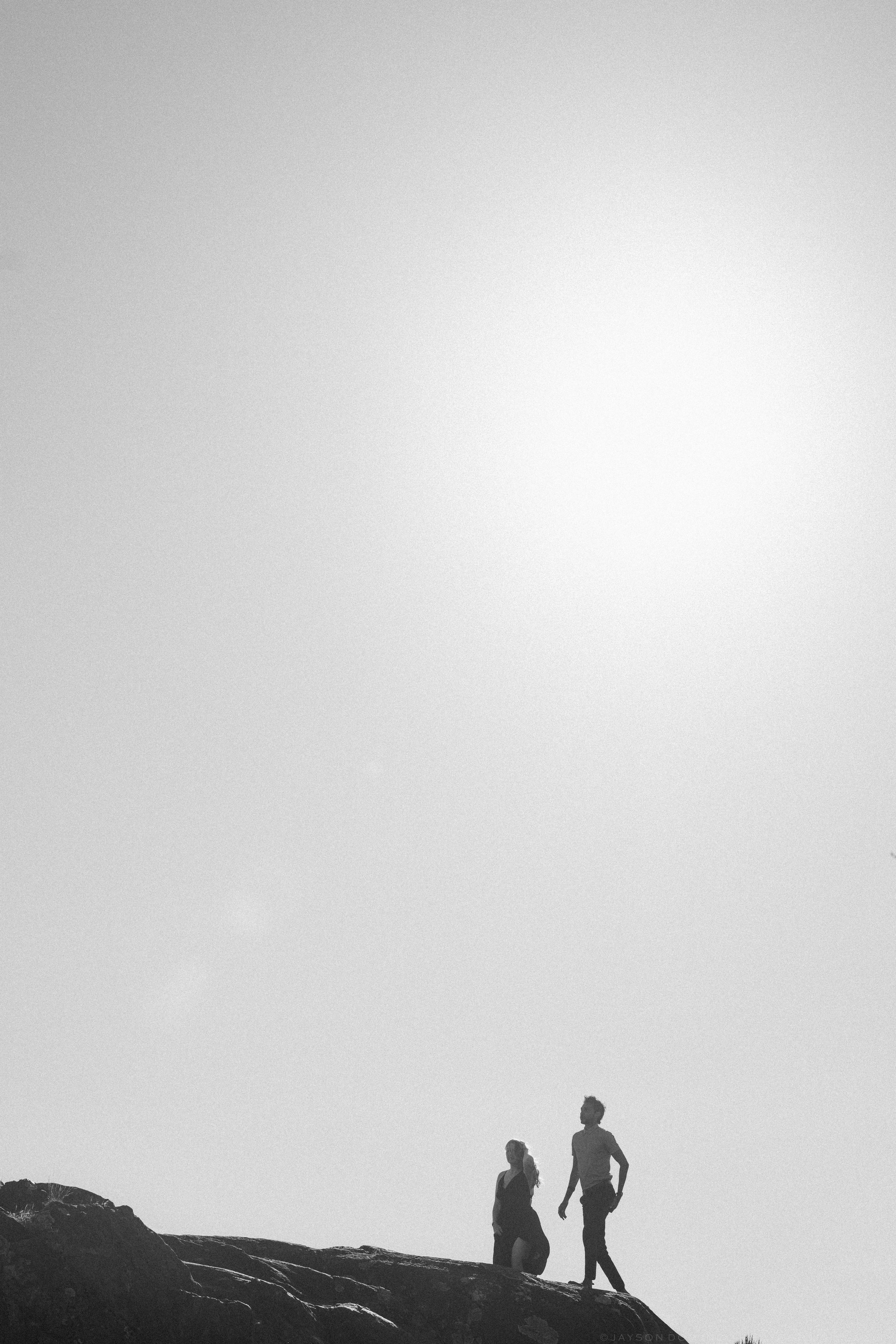 Silhouette of two people standing on rocky terrain against a bright, hazy sky in black and white.