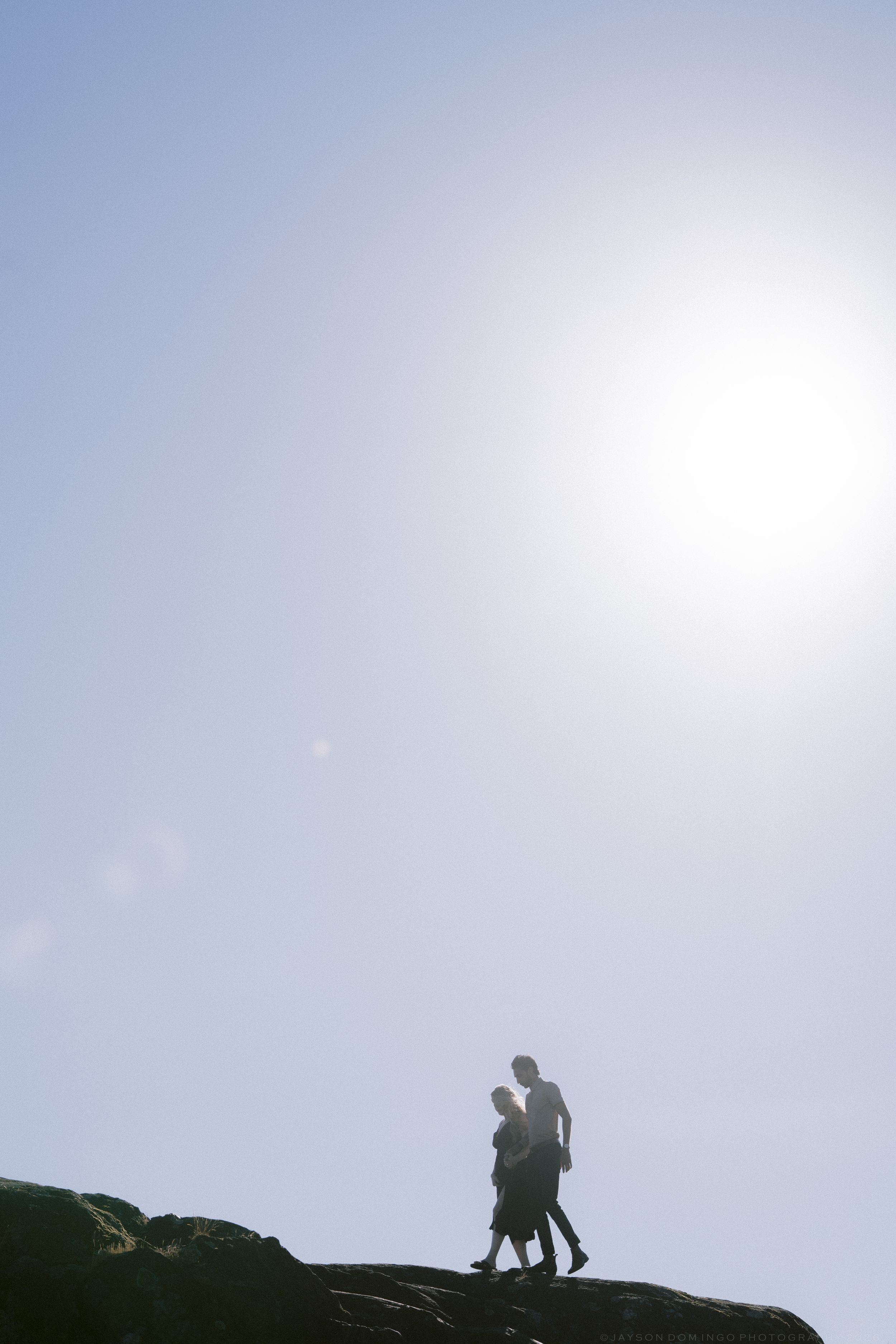 Silhouette of couple walking together on rocky terrain against a bright sunlit sky.