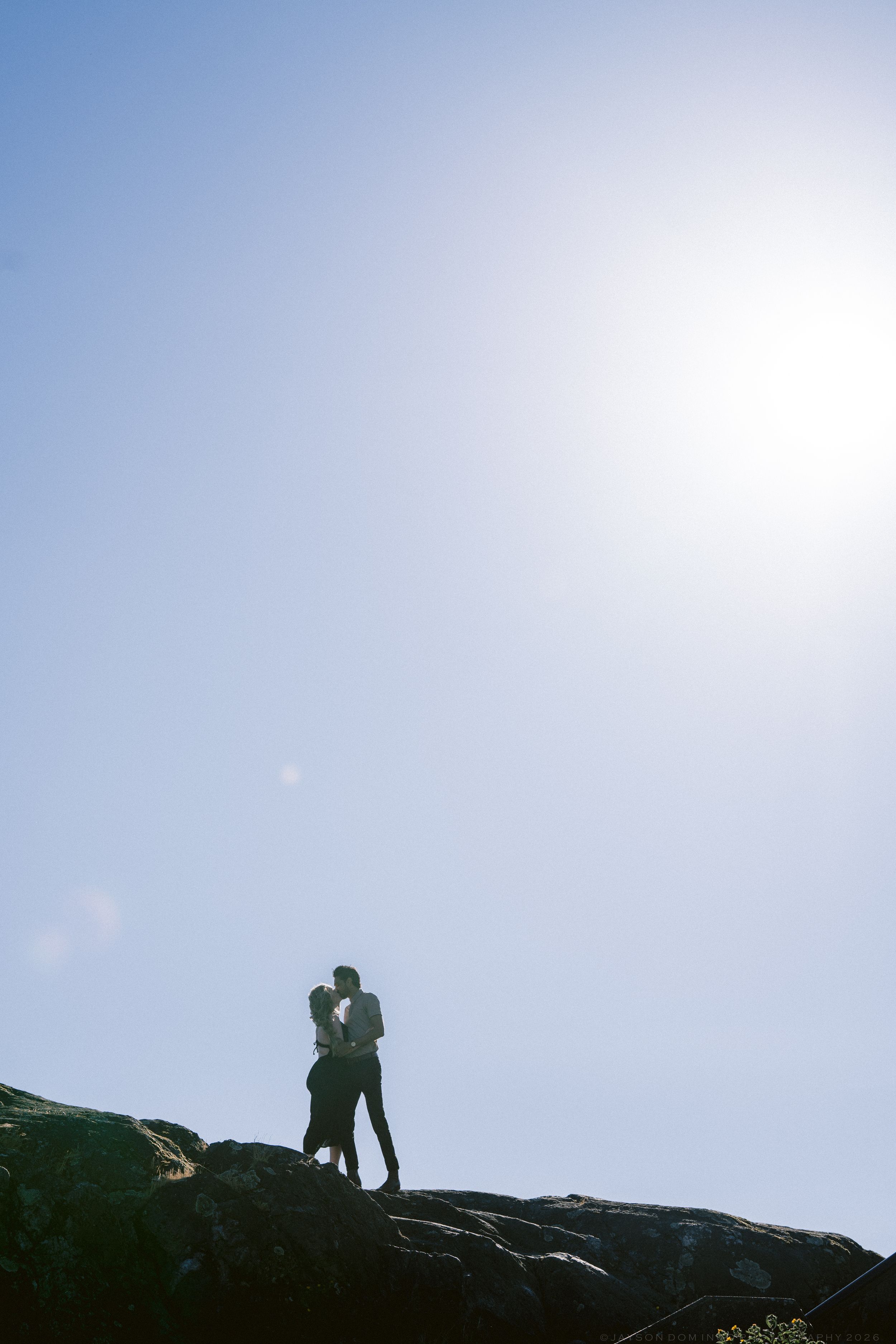 Silhouette of couple embracing on rocky outcrop against bright sunny sky.