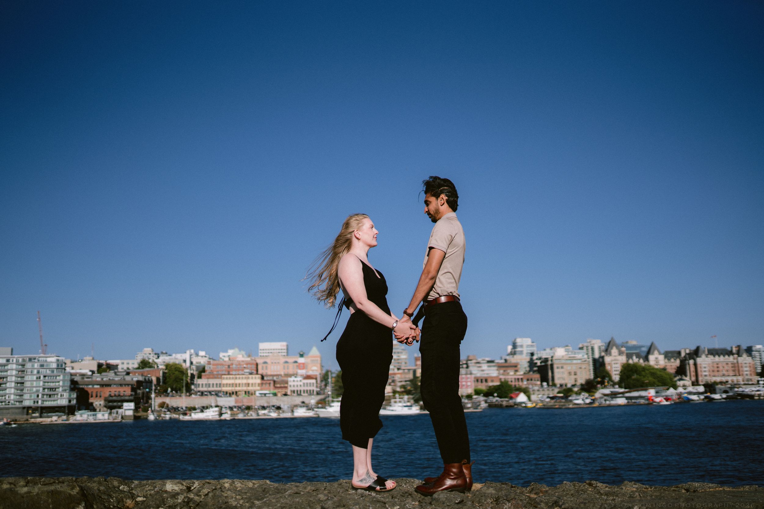 Couple holding hands facing each other by the waterfront with a city skyline and clear blue sky behind them.