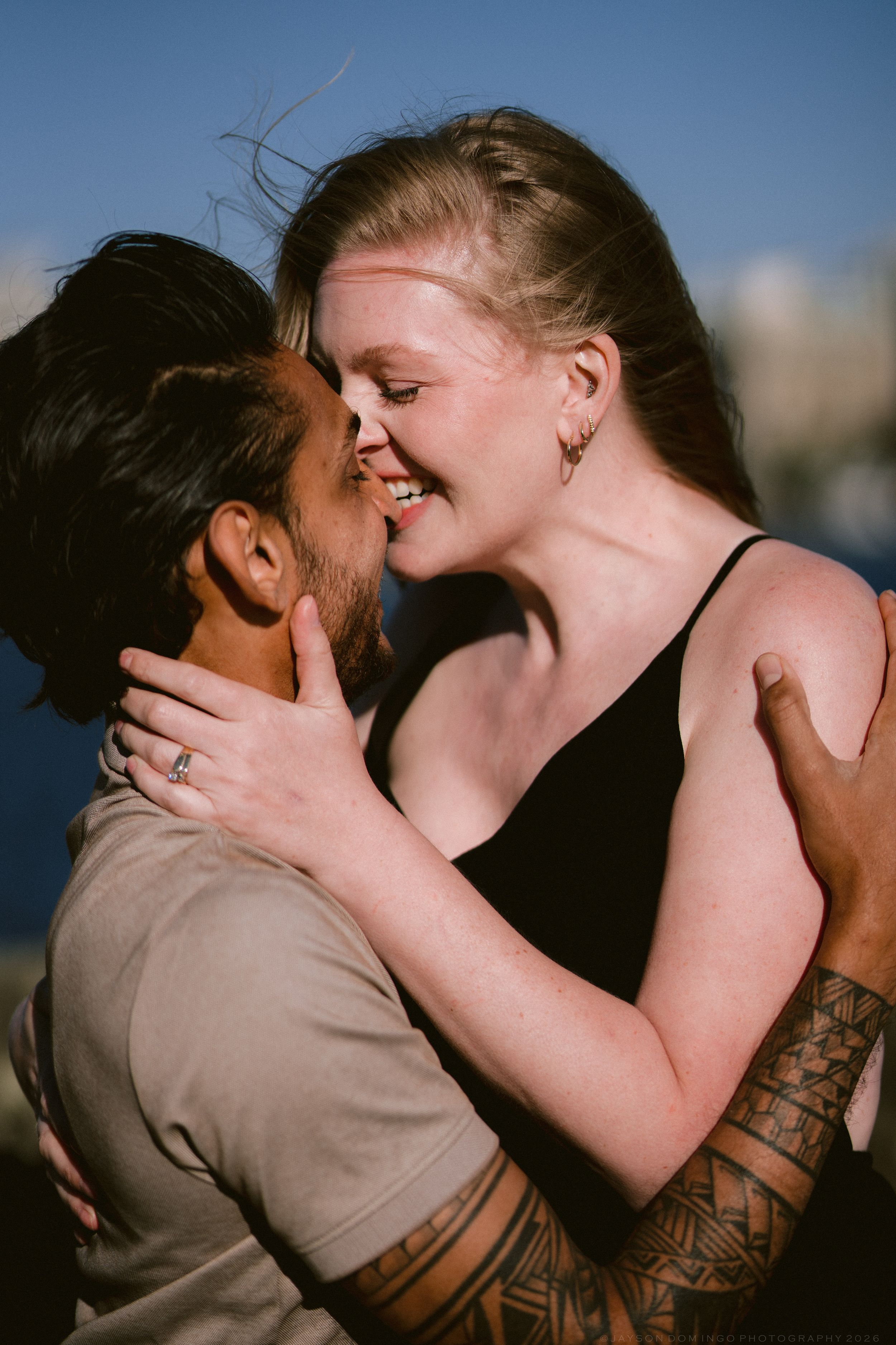 Joyful couple sharing a kiss outdoors with blue sky, woman smiling with tattooed partner.