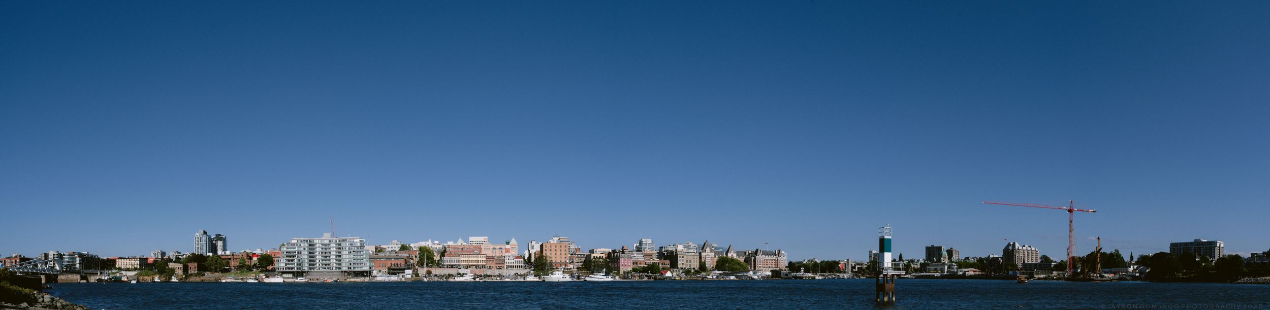 Panoramic skyline view of a coastal city with tall buildings and a tower reflected in calm blue water under a clear sky.