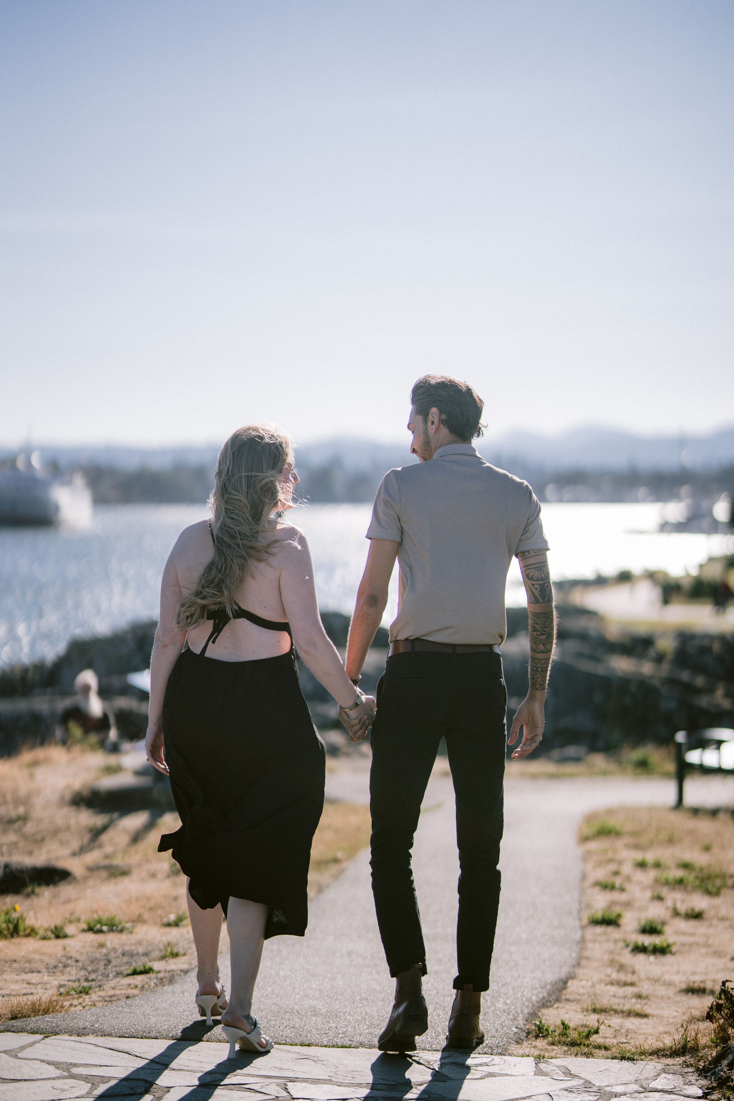 Couple walking hand in hand along sunny waterfront path with ocean and city views behind them.