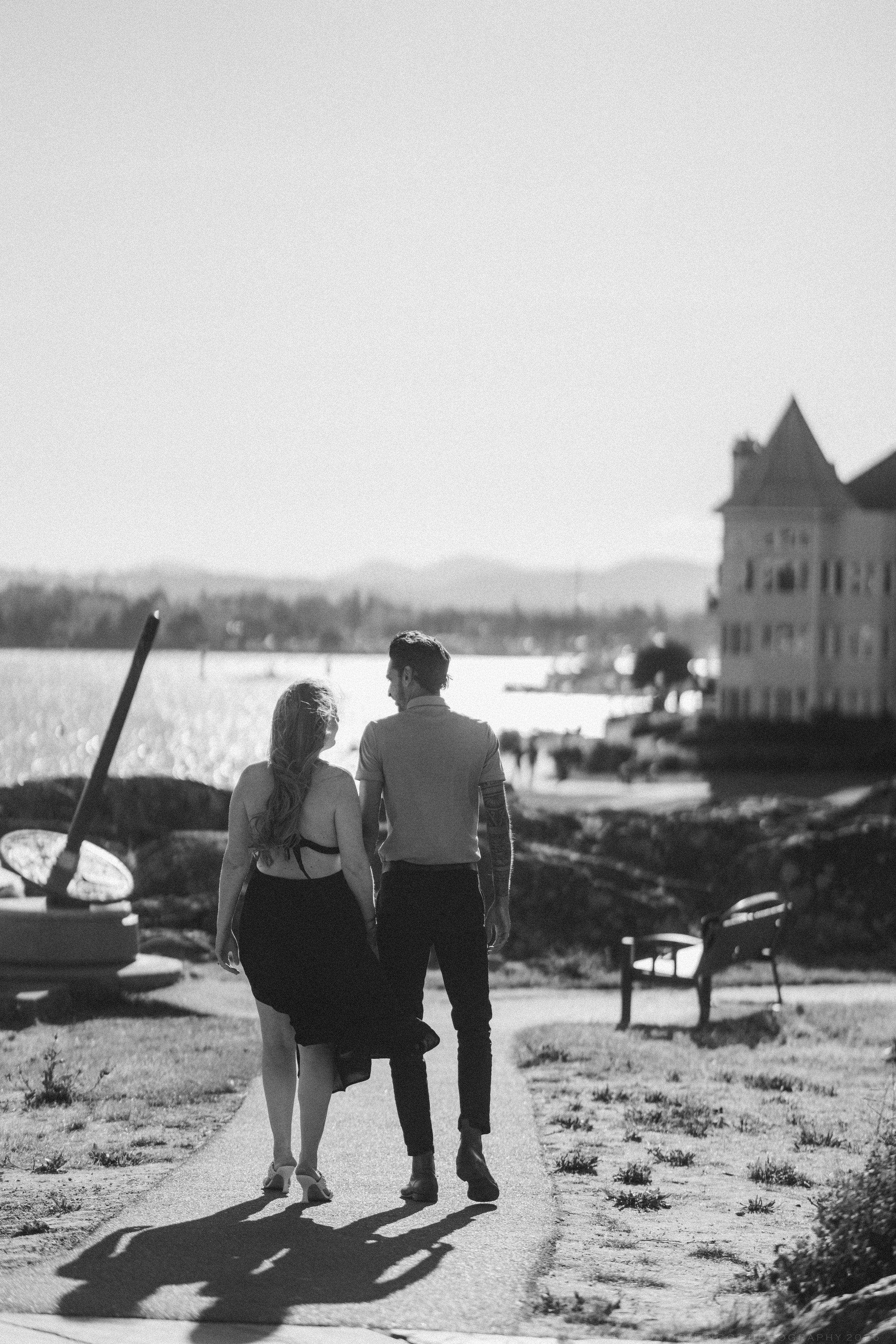 Black and white photo of couple walking hand in hand along waterfront path near historic building.
