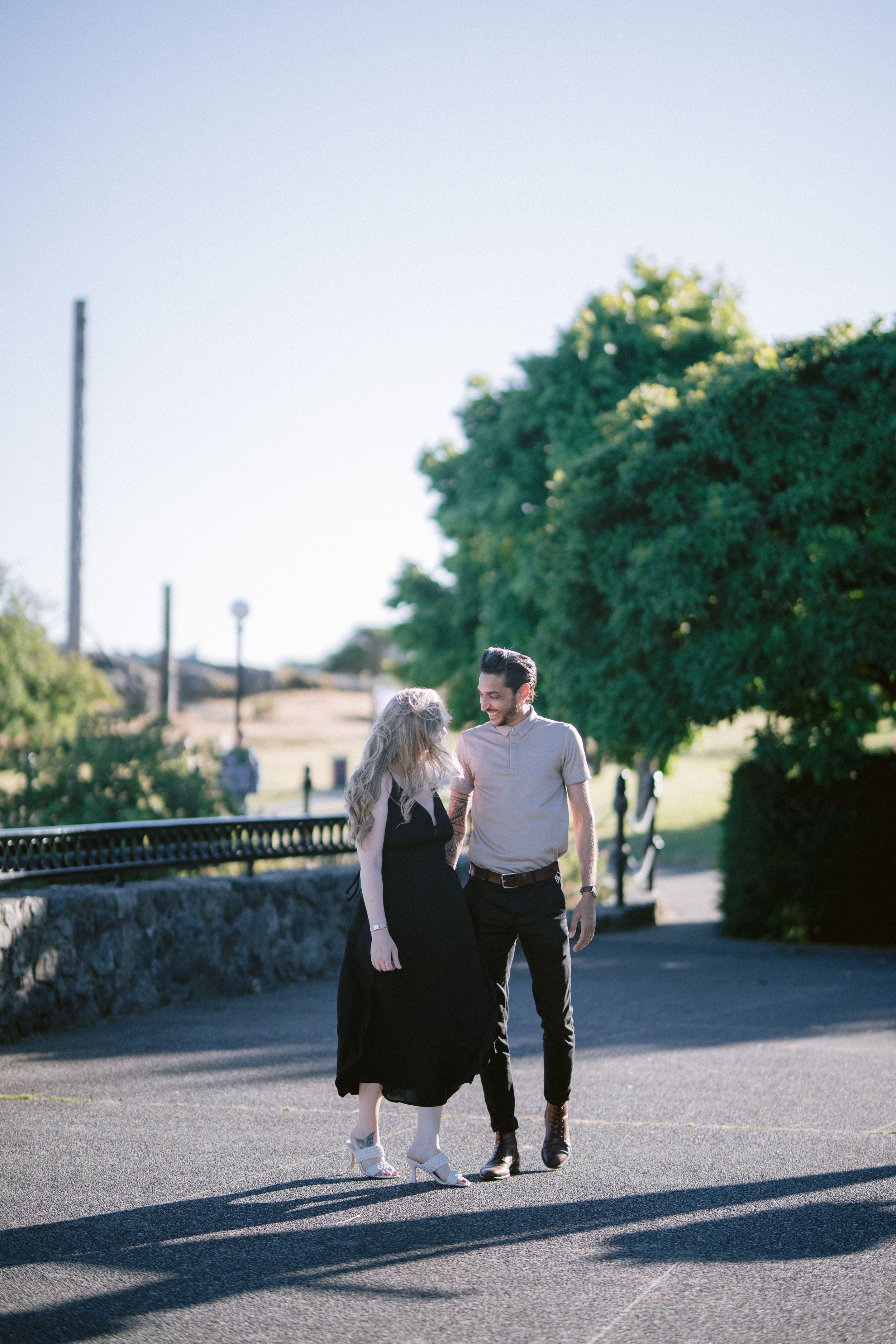 Couple walking together in sunny park surrounded by green trees.