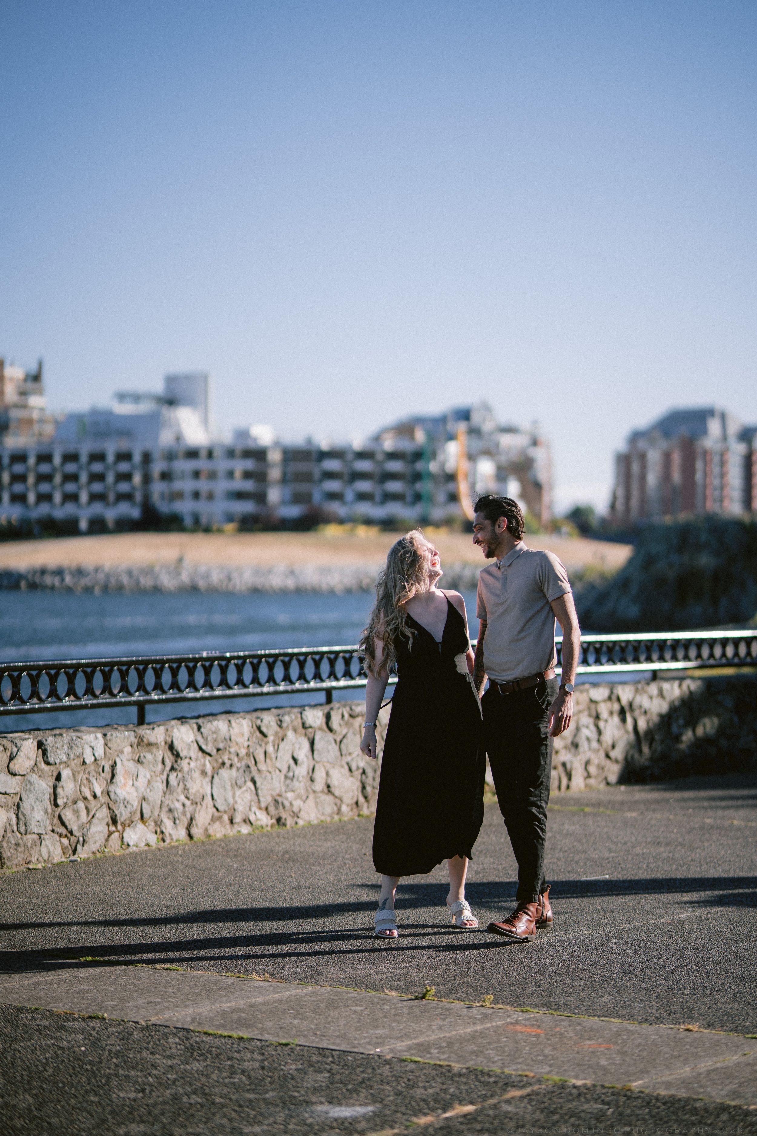 Couple holding hands and walking along a waterfront promenade with city buildings in the background.