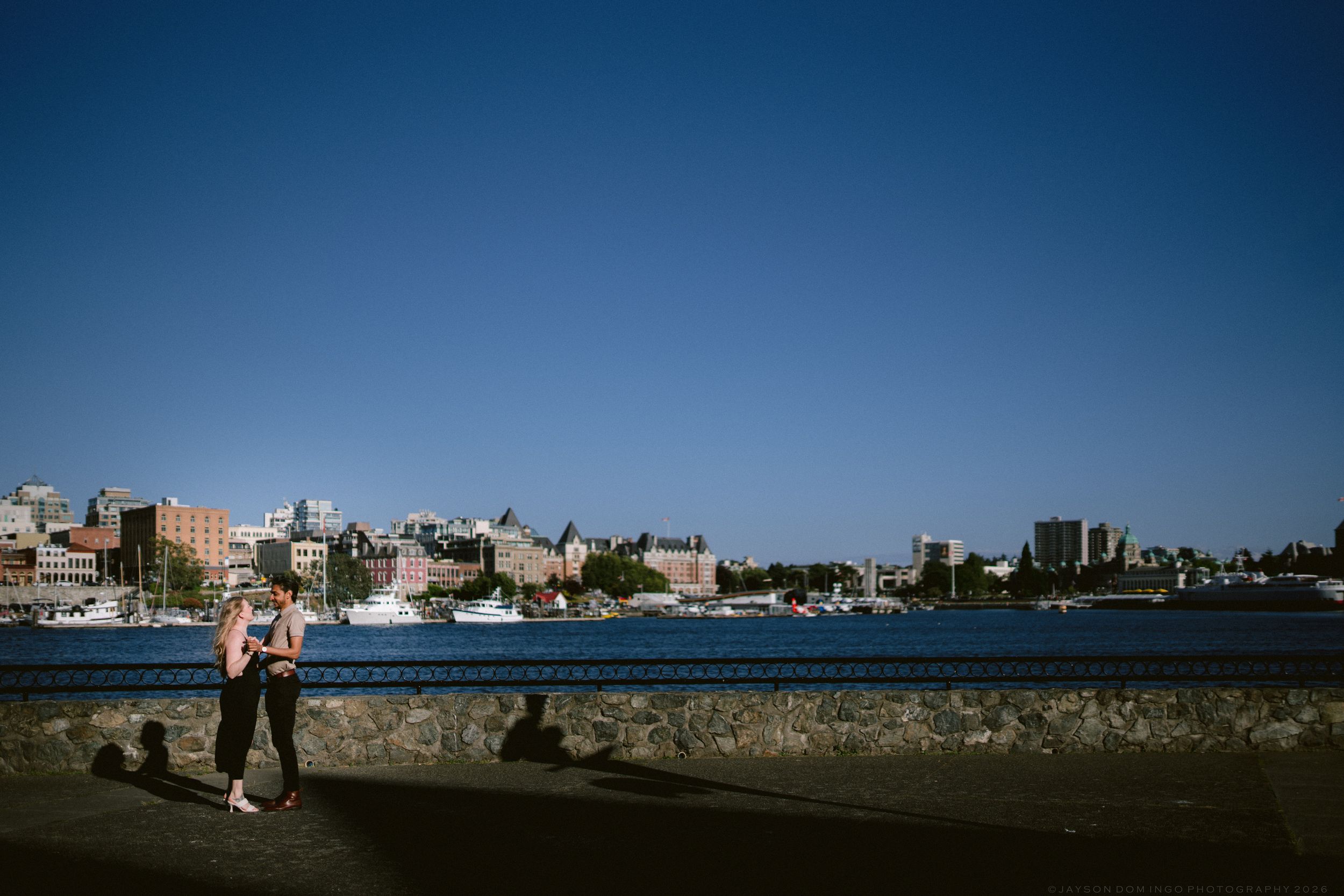 Couple walking along a scenic waterfront promenade with a city skyline visible in the background.
