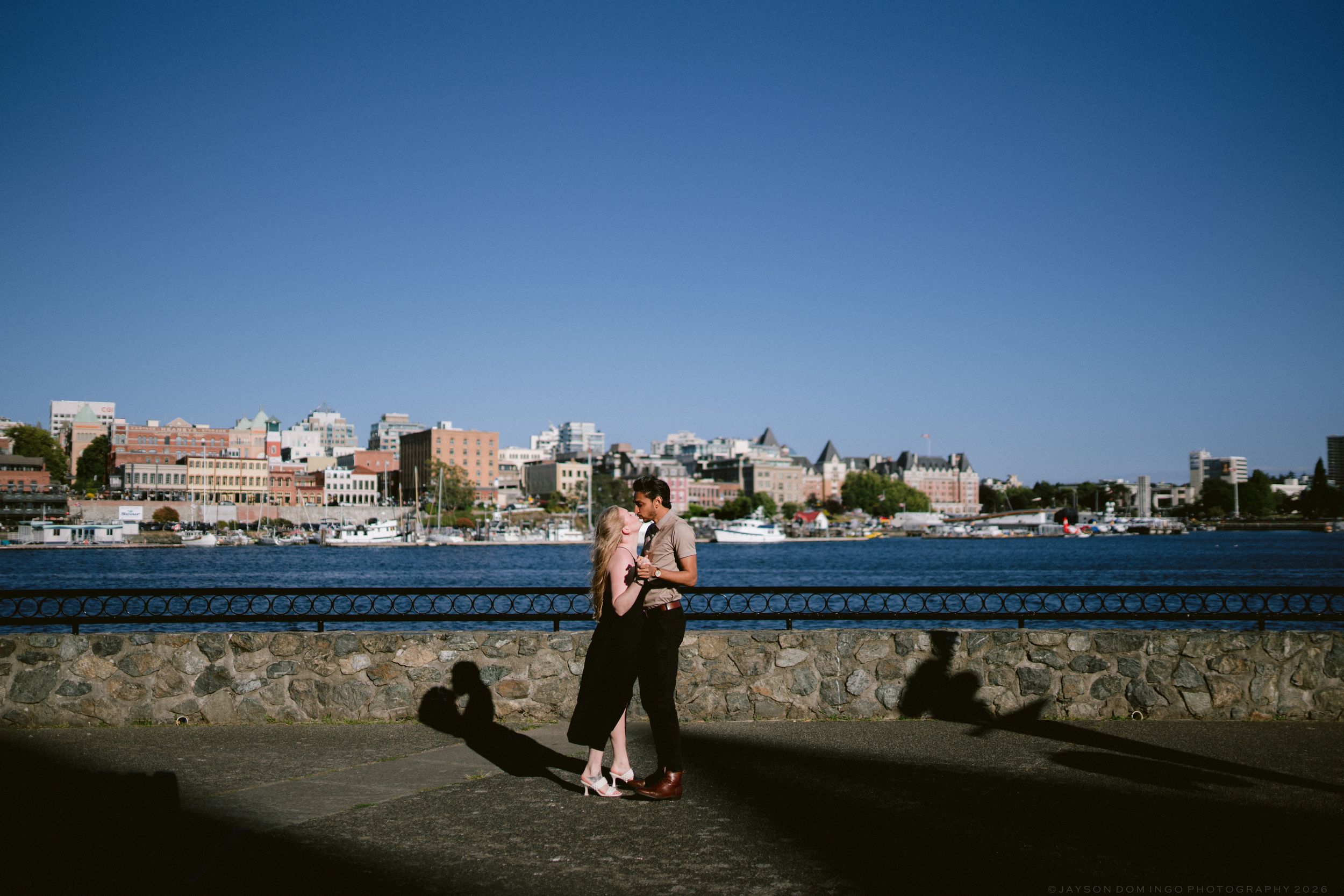 Couple embracing along Victoria BC harbor seawall with blue sky and city skyline backdrop.