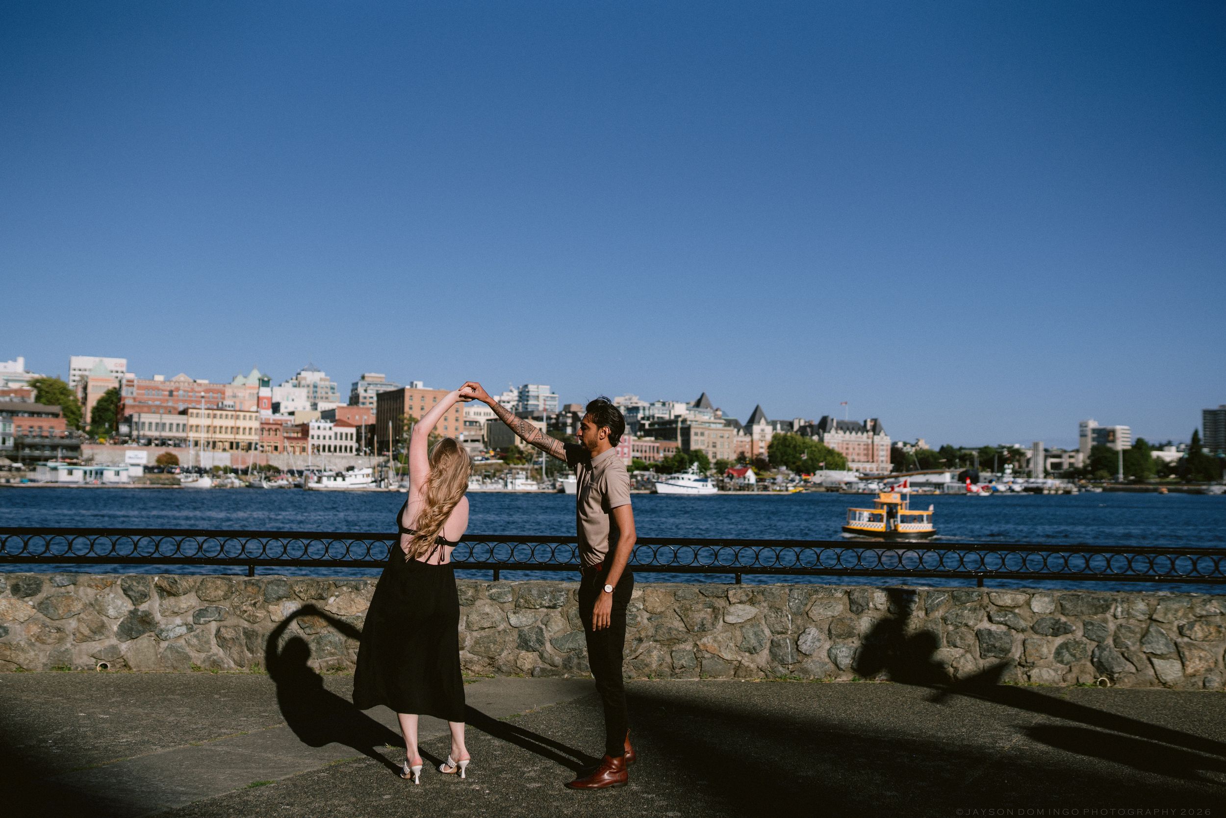 Couple dancing playfully along Victoria BC harbor seawall on a sunny clear day.