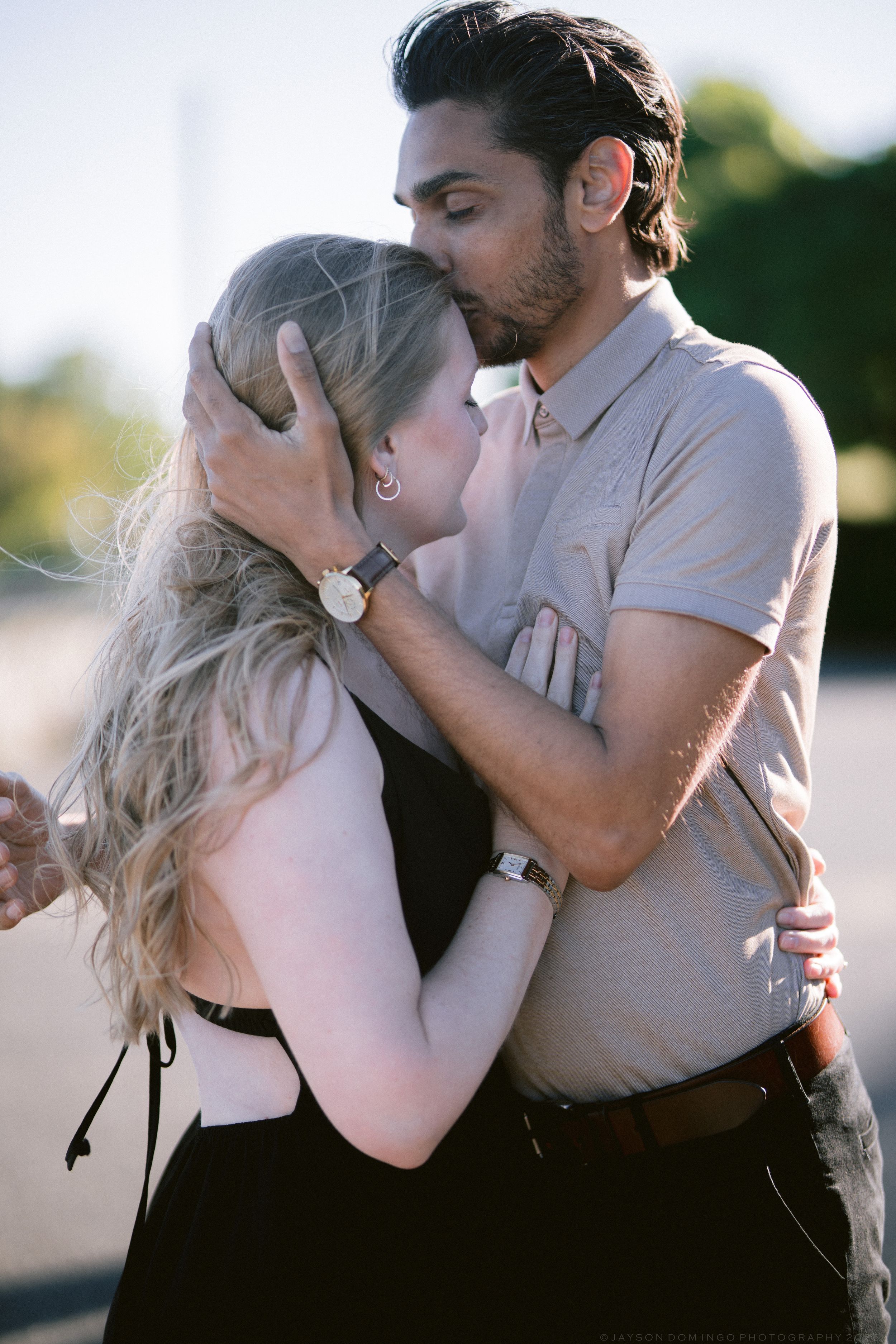 A couple shares a tender kiss outdoors, the man embracing the woman with both hands in warm sunlight.