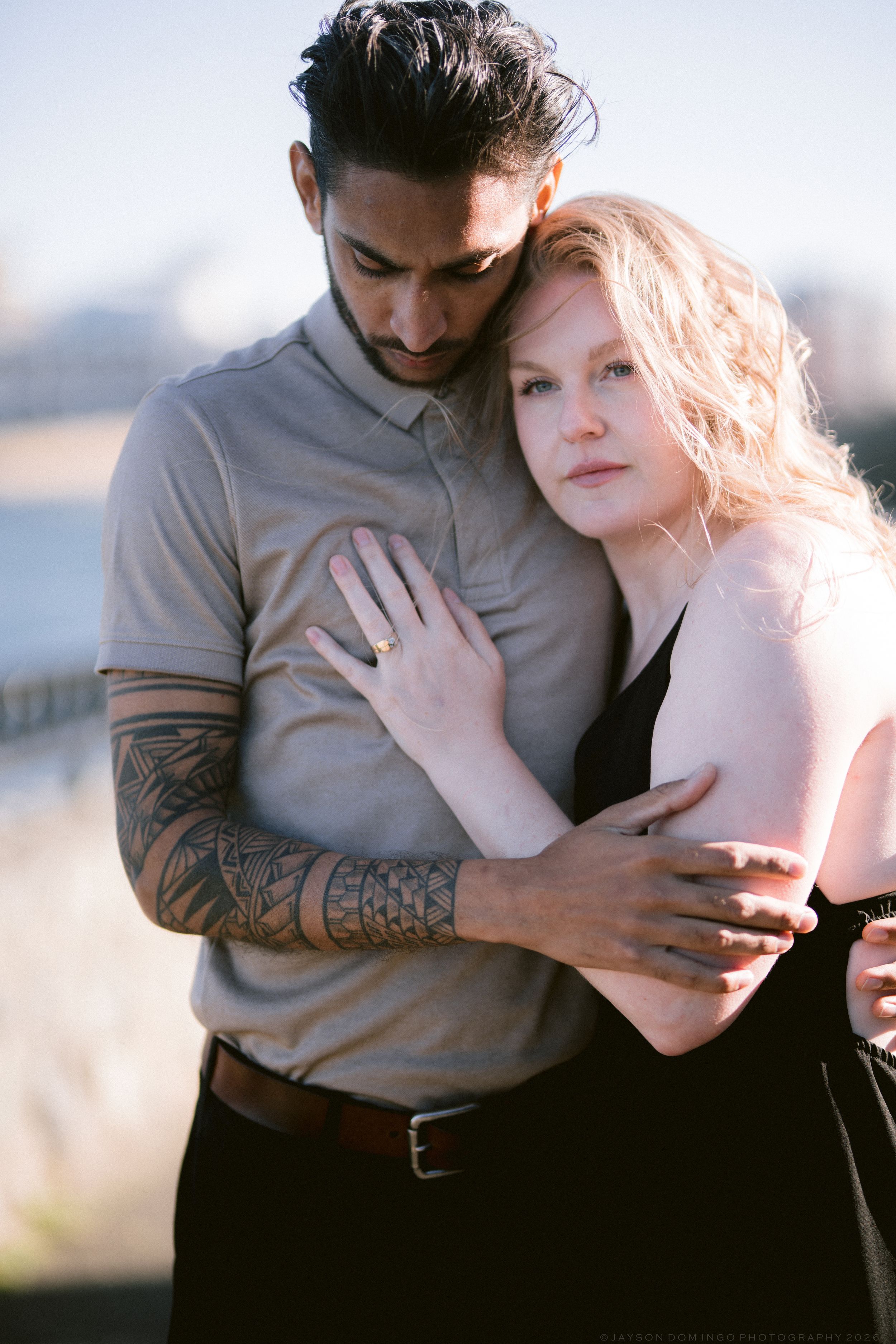 Tattooed man in beige polo embracing blonde woman tenderly outdoors near waterfront in soft light.