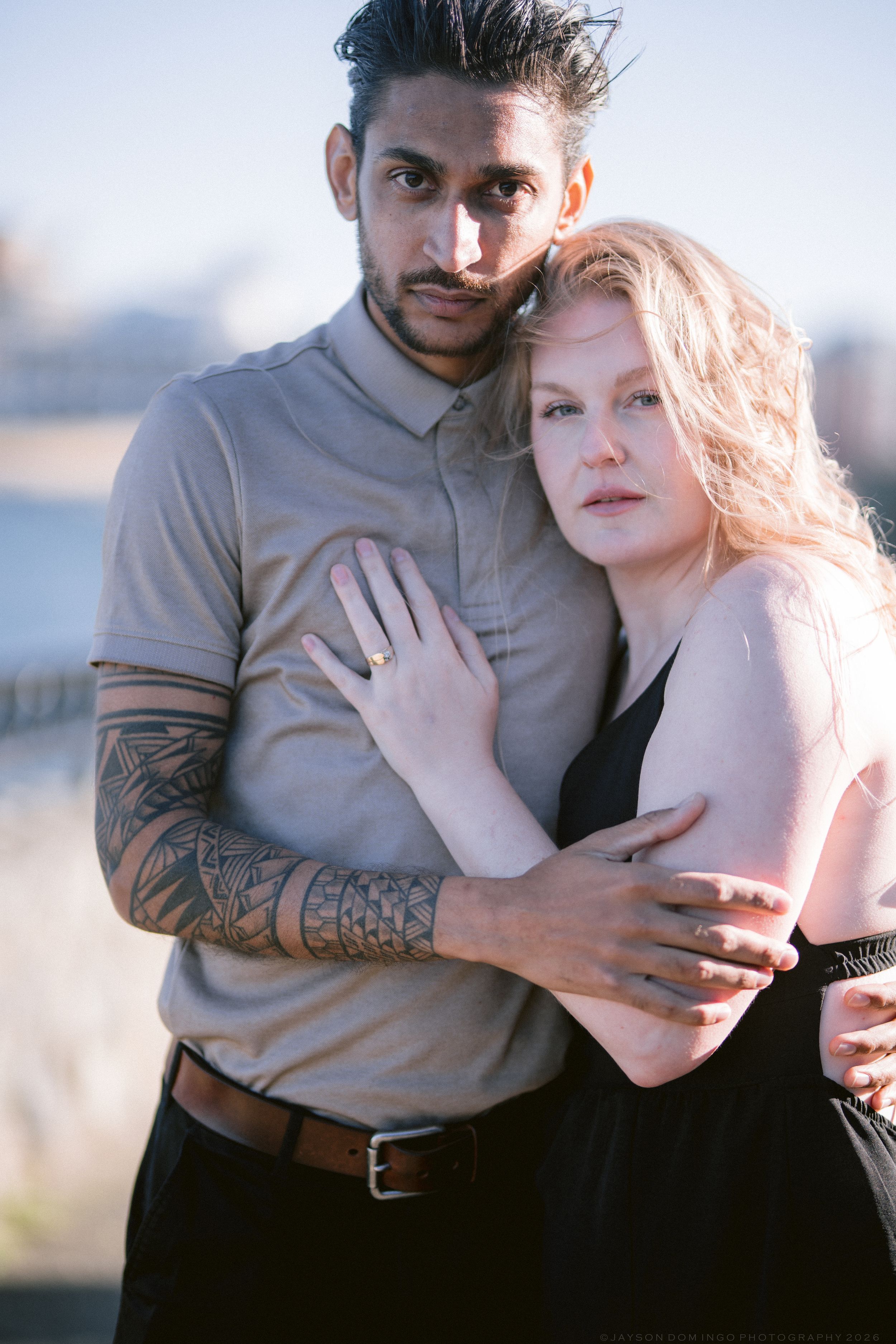 Interracial couple embracing outdoors, man with sleeve tattoo holding woman in black dress.