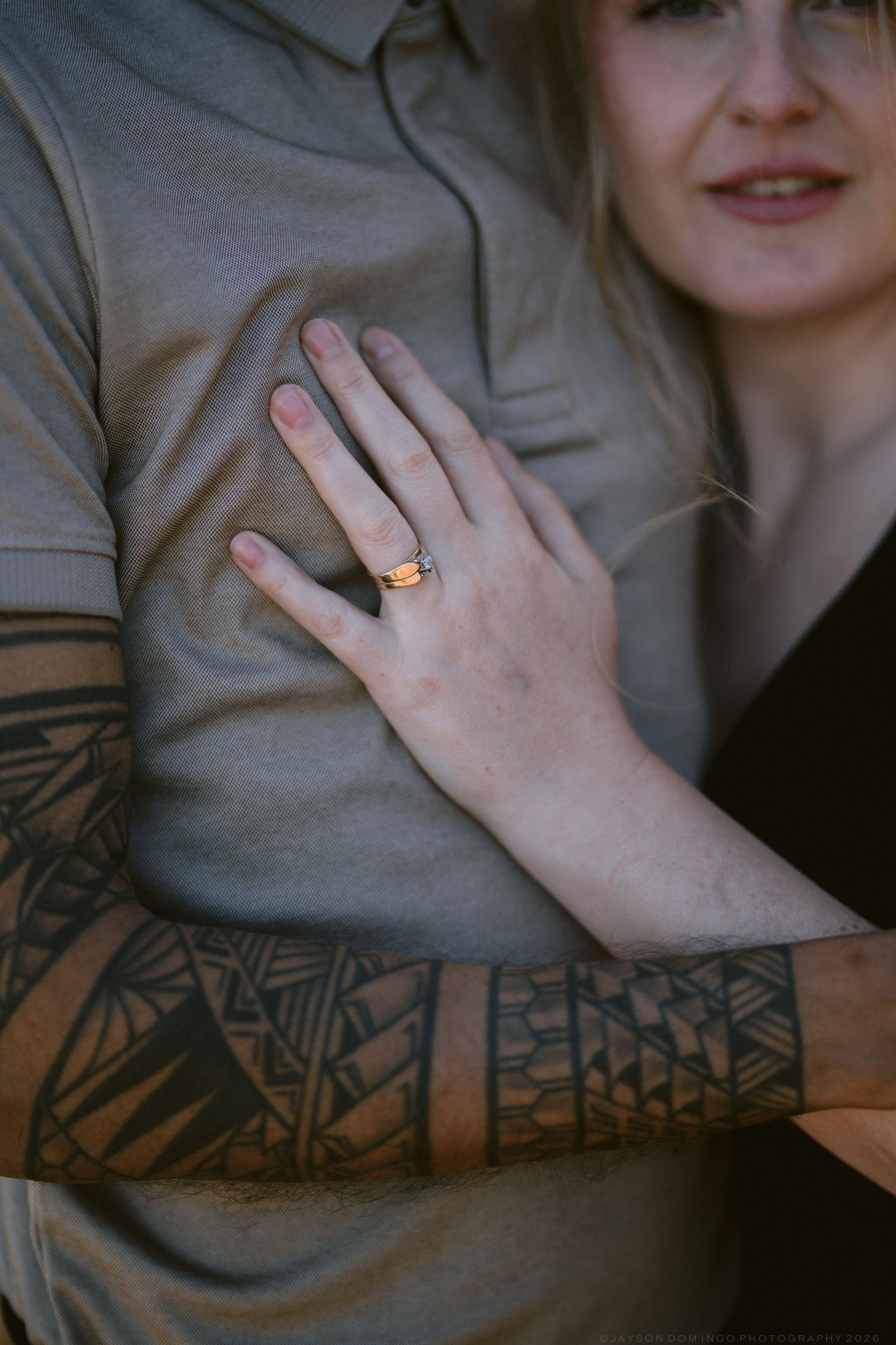 Close-up of a womans hand displaying an engagement ring resting on a mans chest with a tribal tattoo sleeve.