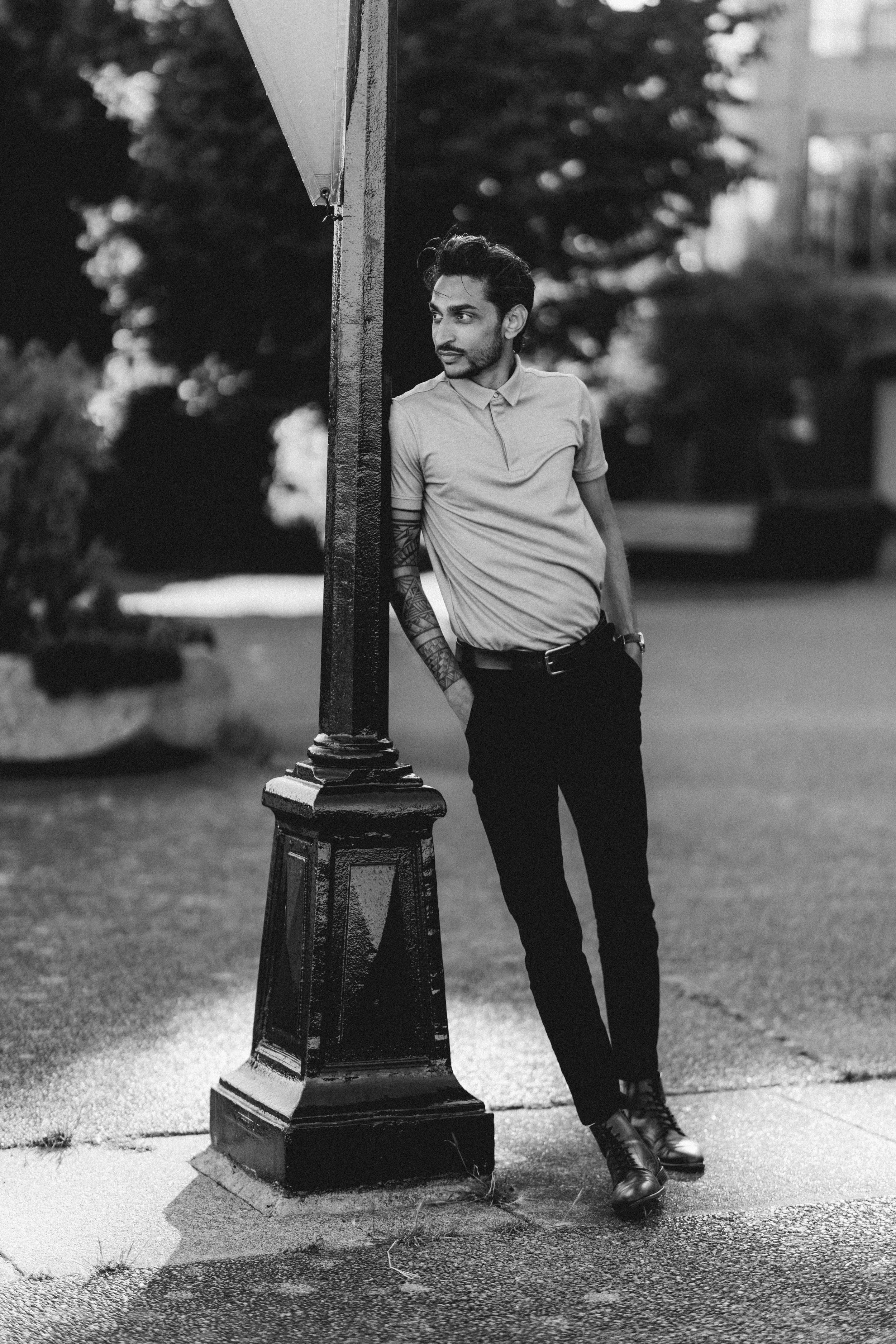 Black and white portrait of a tattooed man leaning against a lamppost in an urban setting.