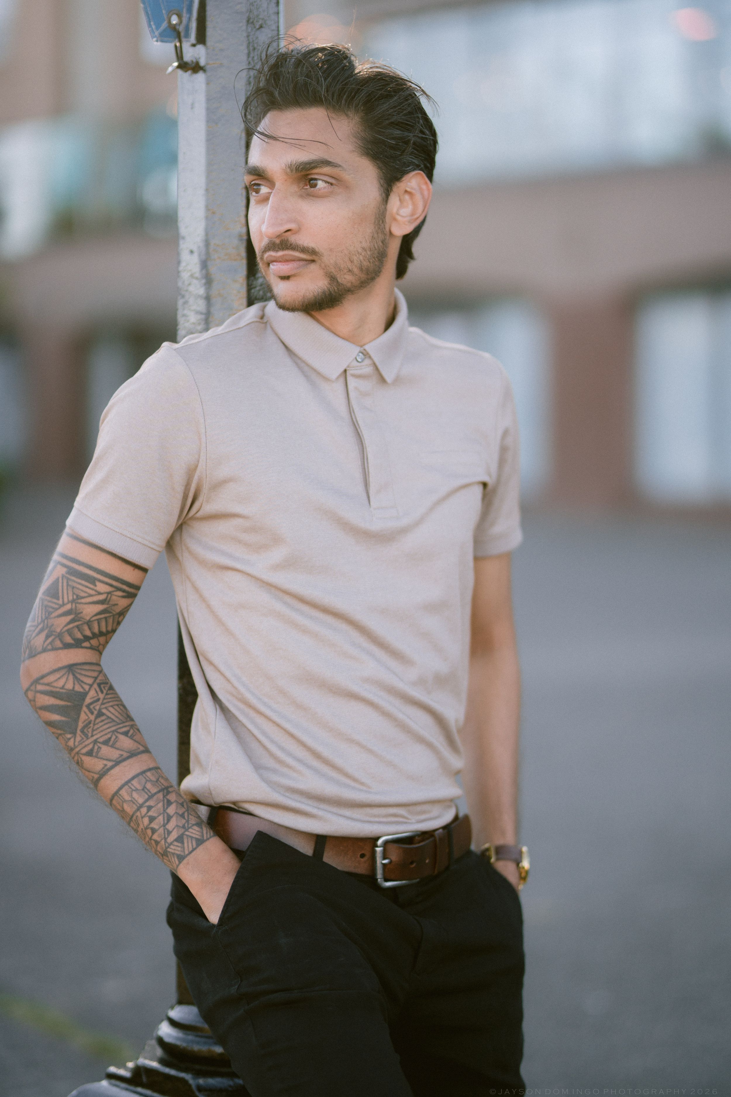 Tattooed man in beige polo shirt leaning against pole in urban setting.